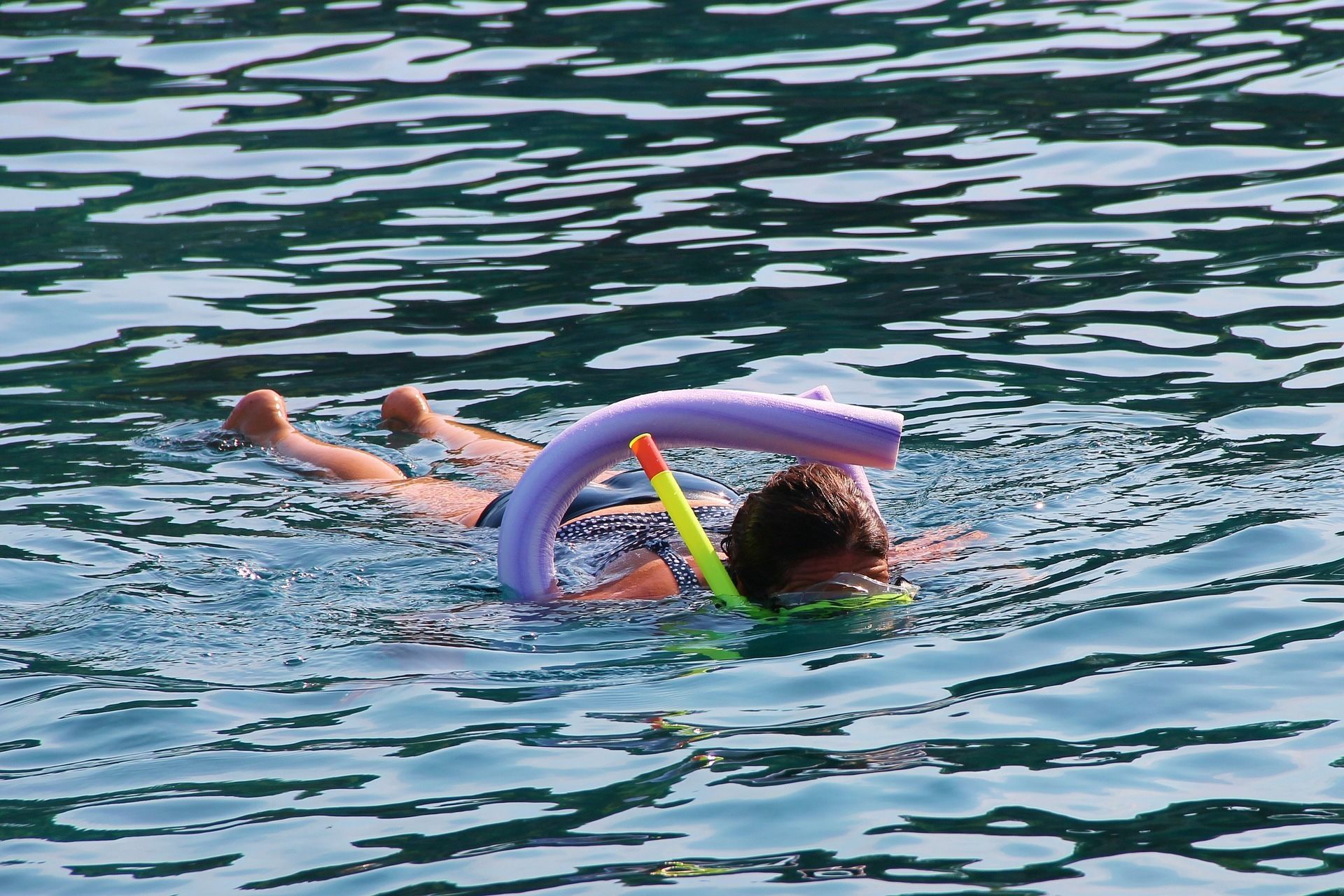 Child snorkeling with a purple pool noodle, floating in blue water.