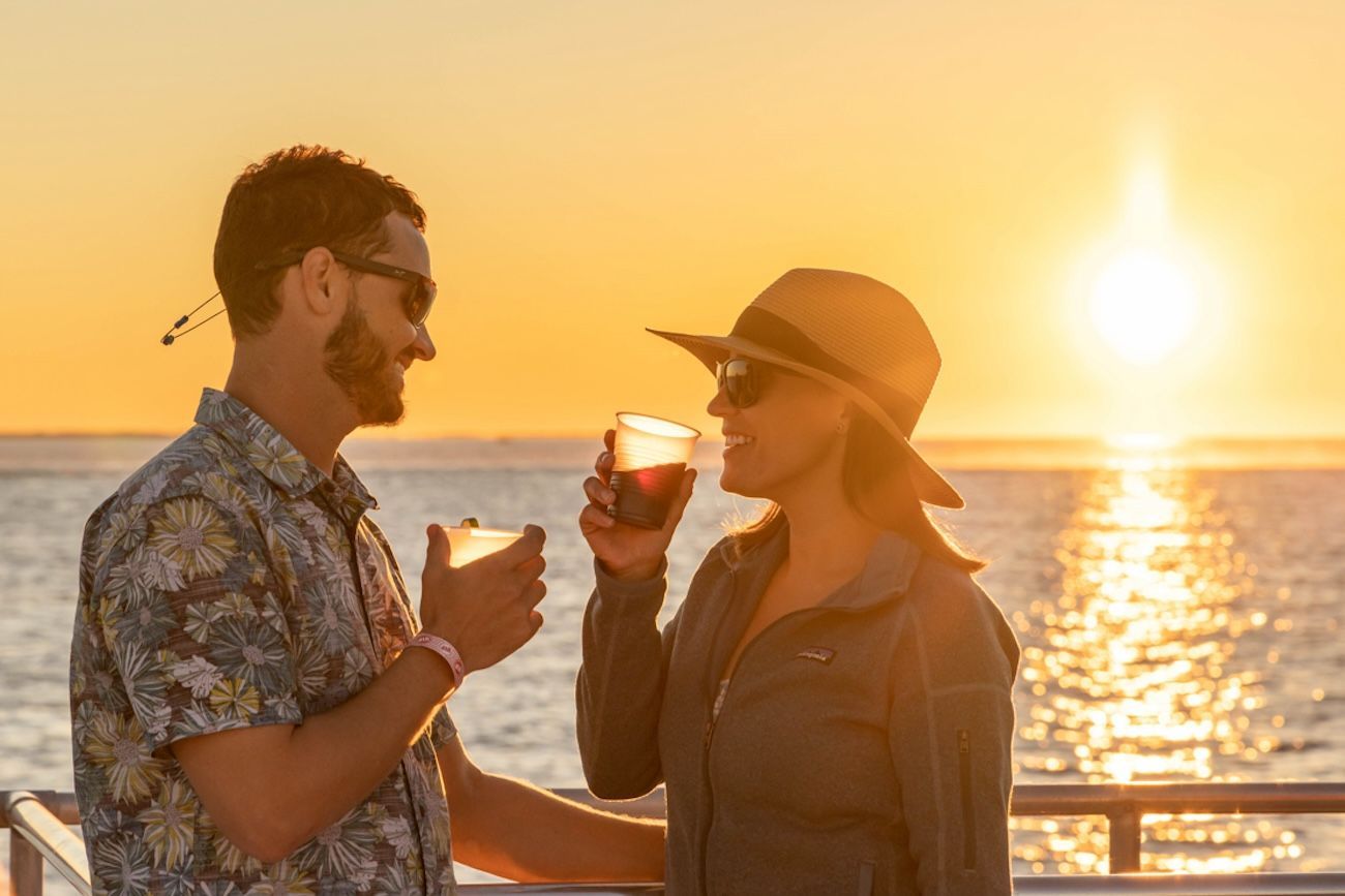 Couple toasting drinks, smiling, at sunset over the ocean.