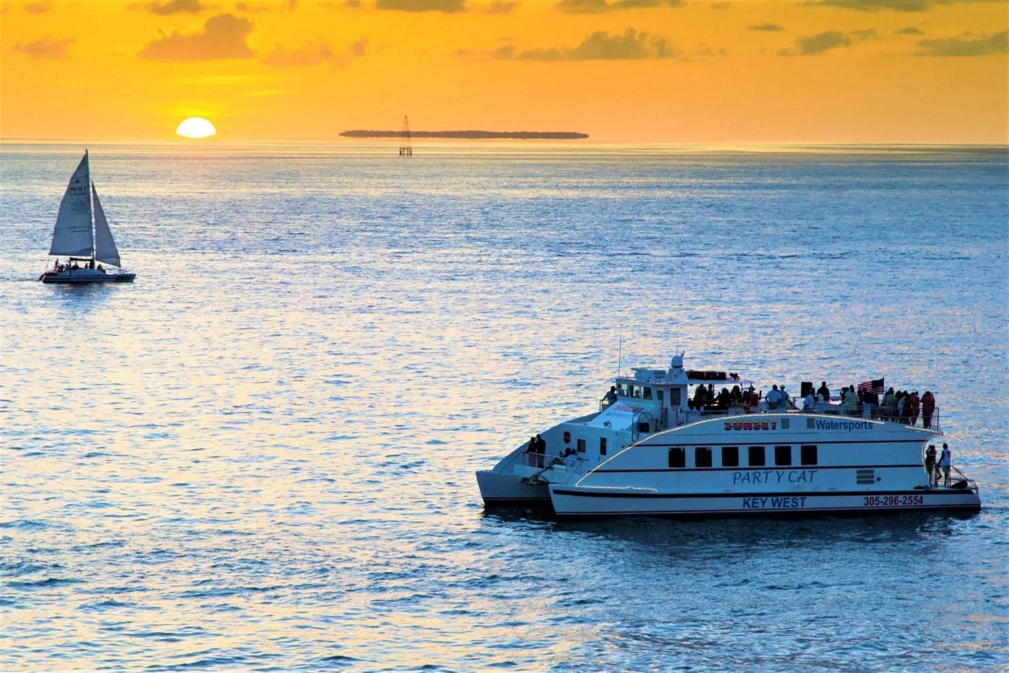 Sunset over the ocean with a sailboat and a catamaran filled with people.