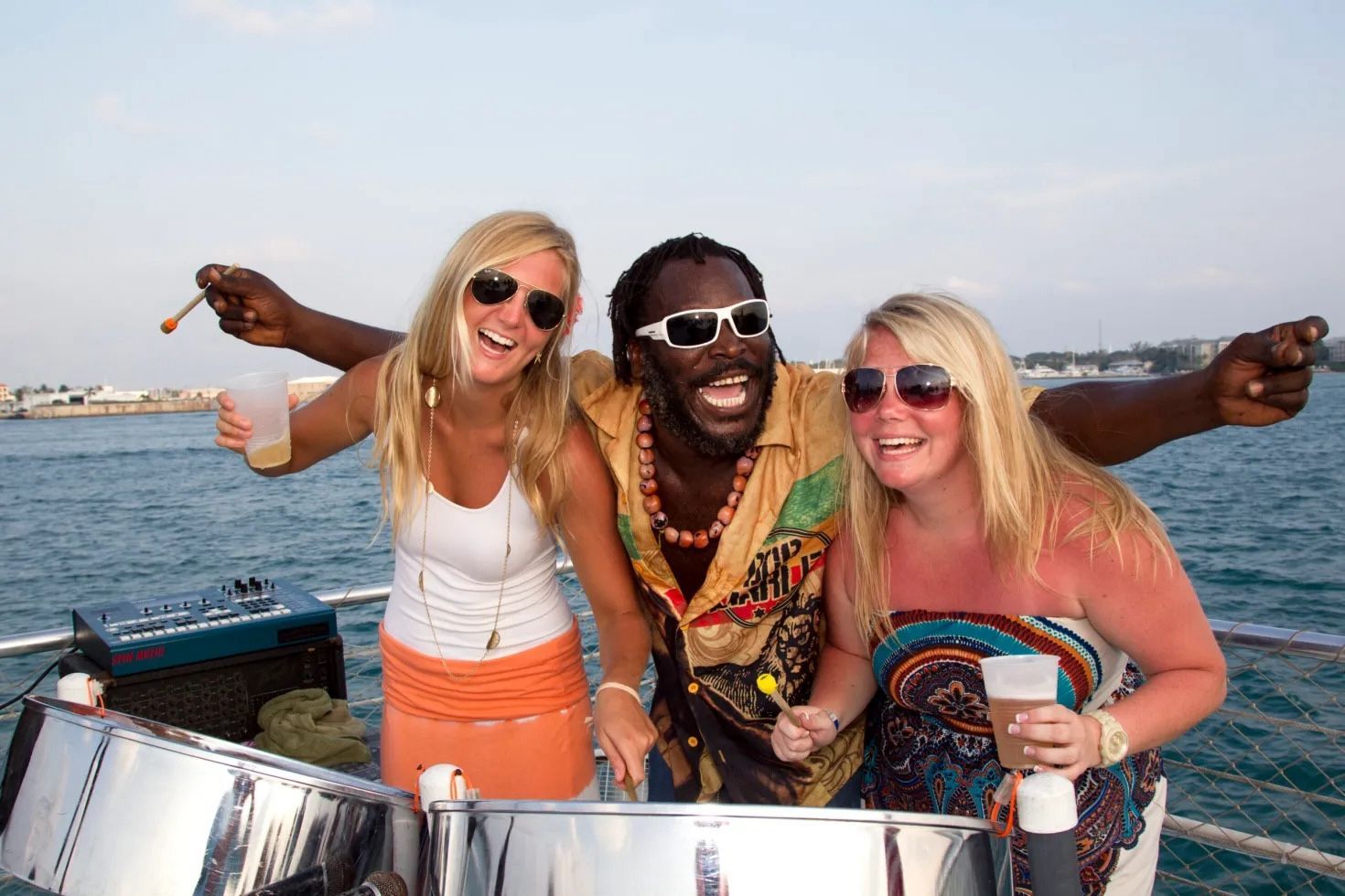 Three people on a boat: a man playing steel drums, flanked by two smiling women holding drinks, sea in background.