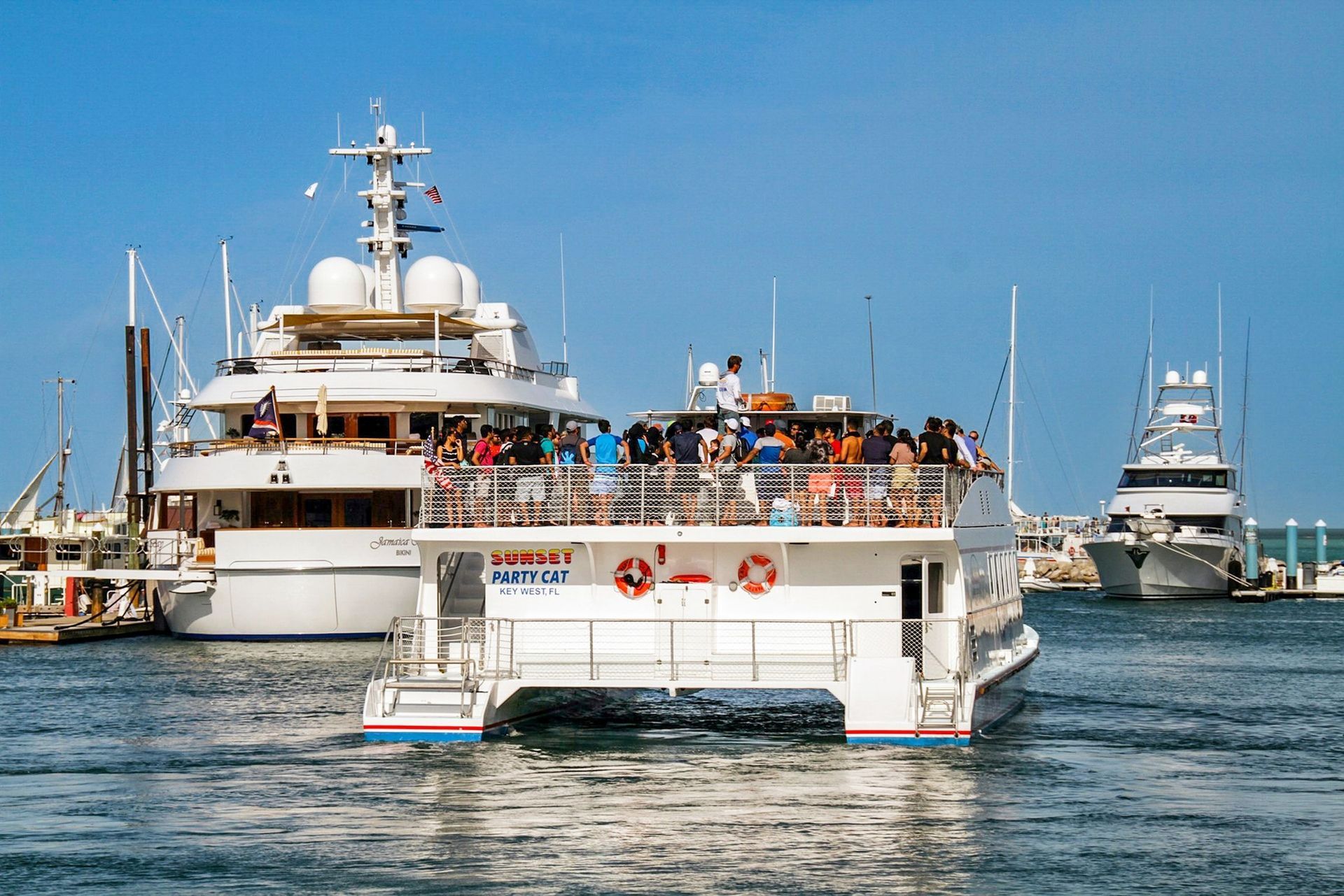 Boats on a harbor. A large party boat filled with people passes a docked white yacht on a sunny day.