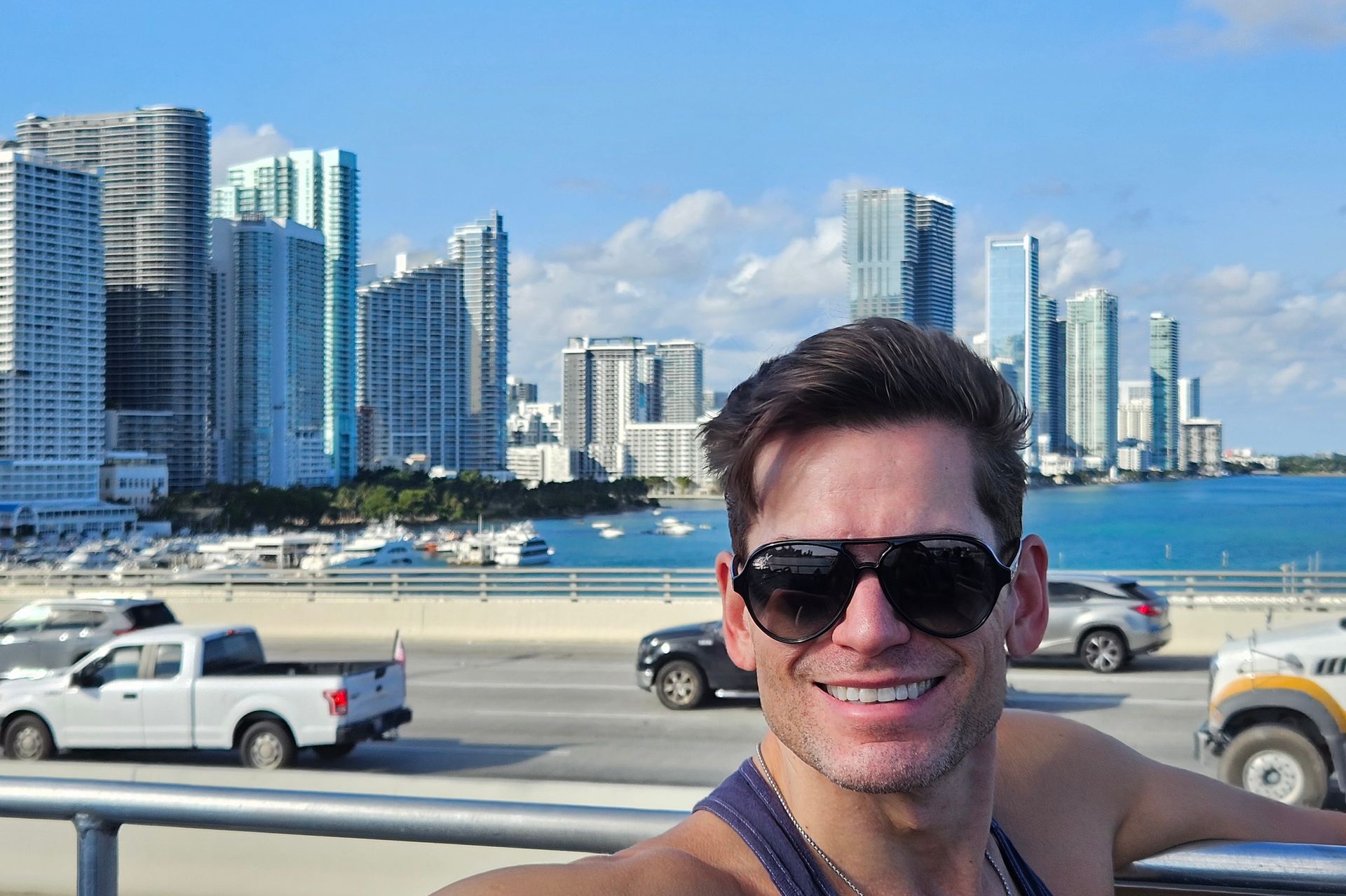 Man smiling, wearing sunglasses, taking a selfie with Miami skyline in the background. Sunny day, buildings, water, vehicles.