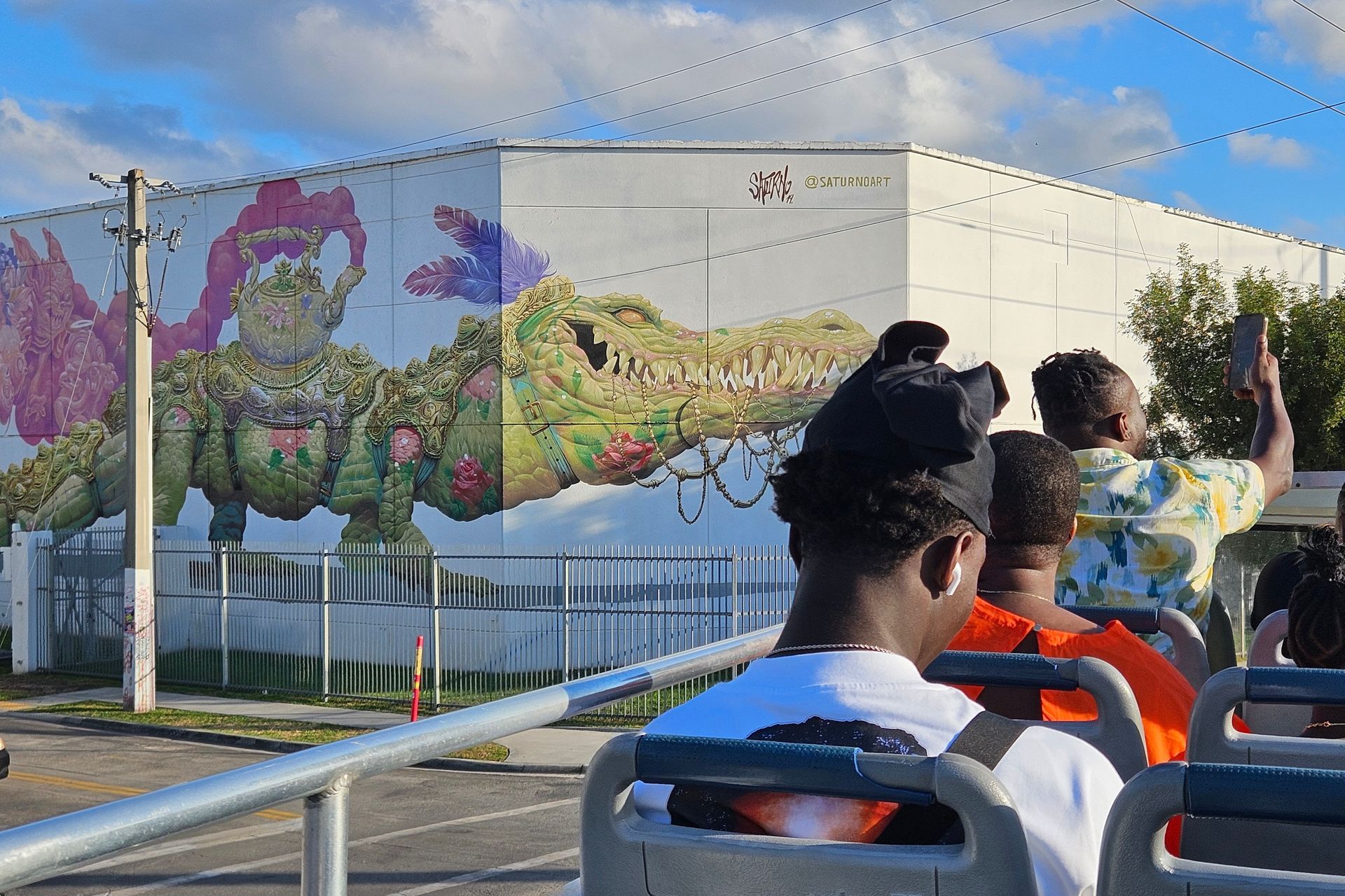 People on a bus taking photos of a colorful mural of an alligator and a character on a white building.