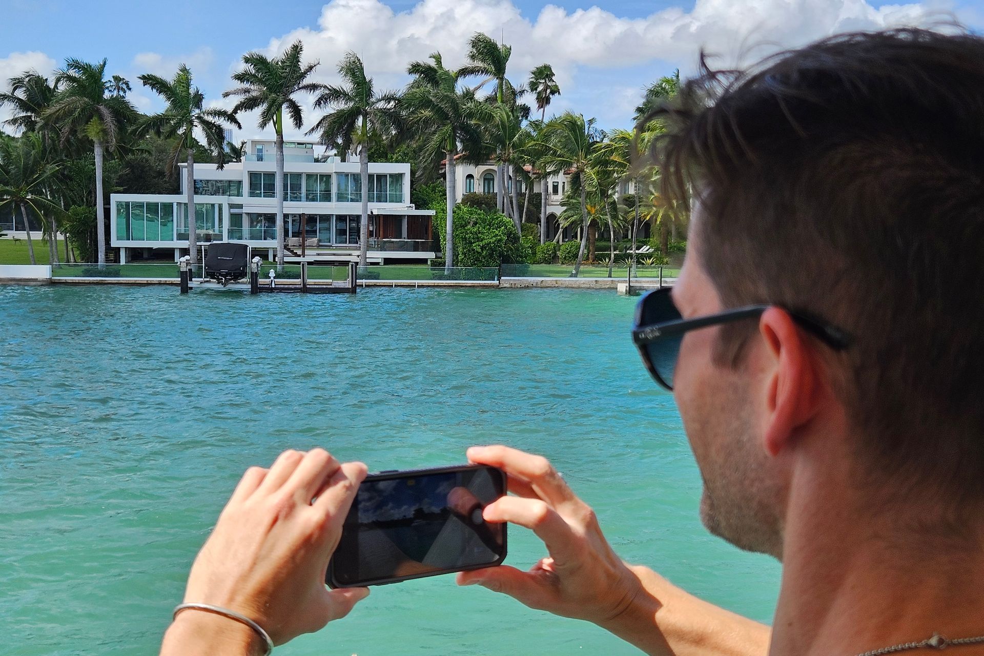 Man taking a photo with his phone of a modern white house by turquoise water, palm trees in background.