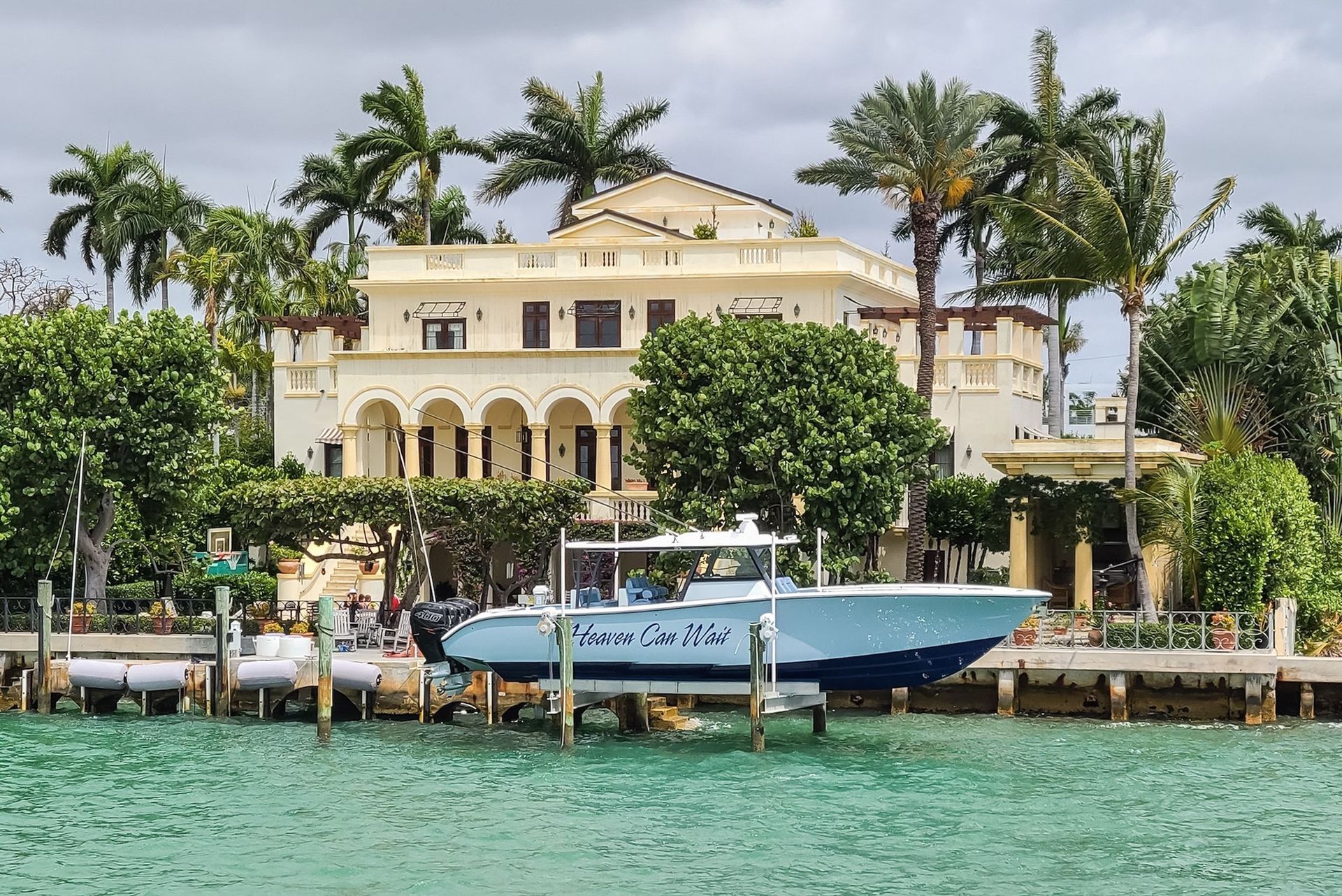 Large beige mansion with boat dock, turquoise water, palm trees, and a blue boat labeled 