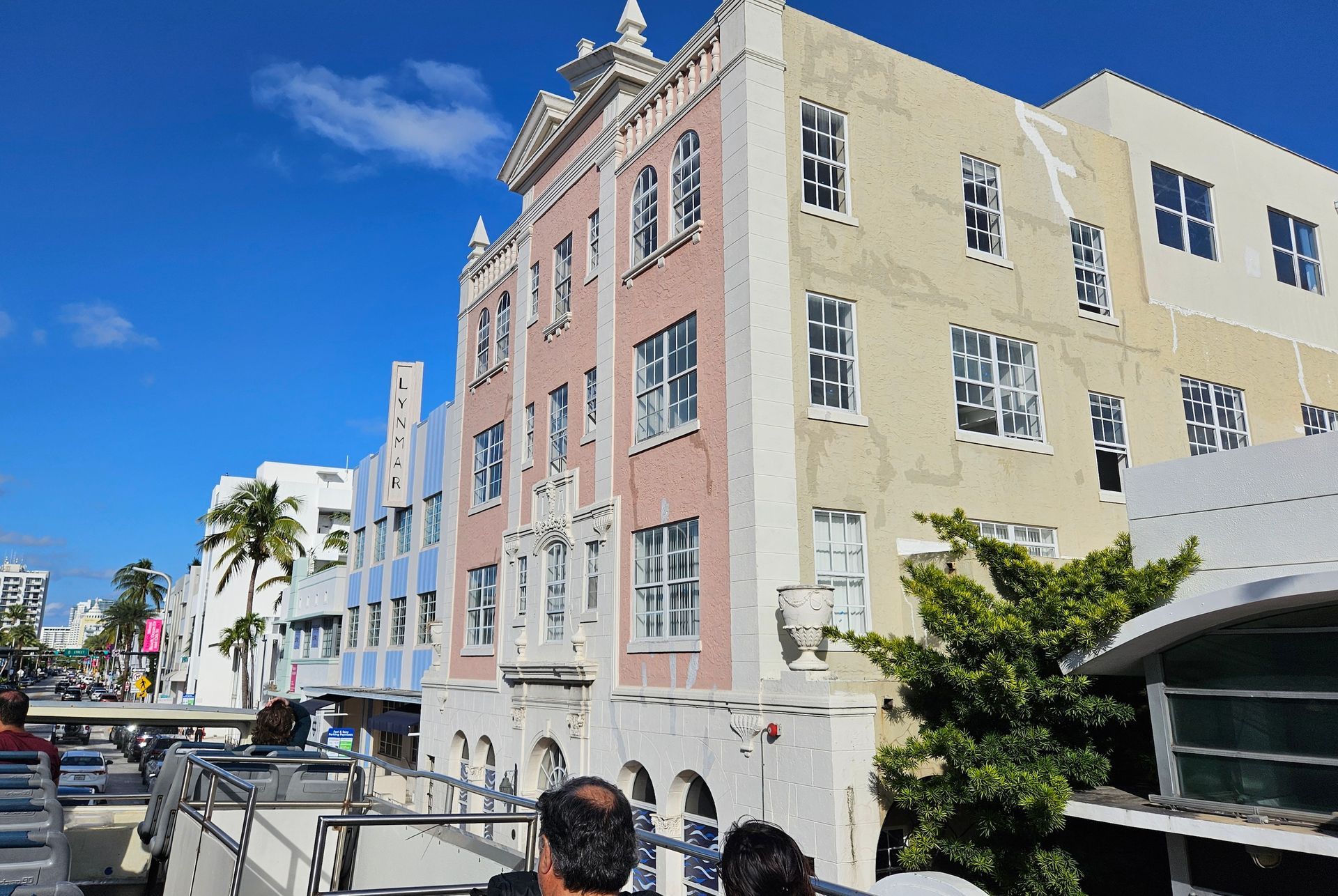 Multi-story buildings in Miami, pink, tan, and white, with arched windows. Clear blue sky, people on a tour bus.