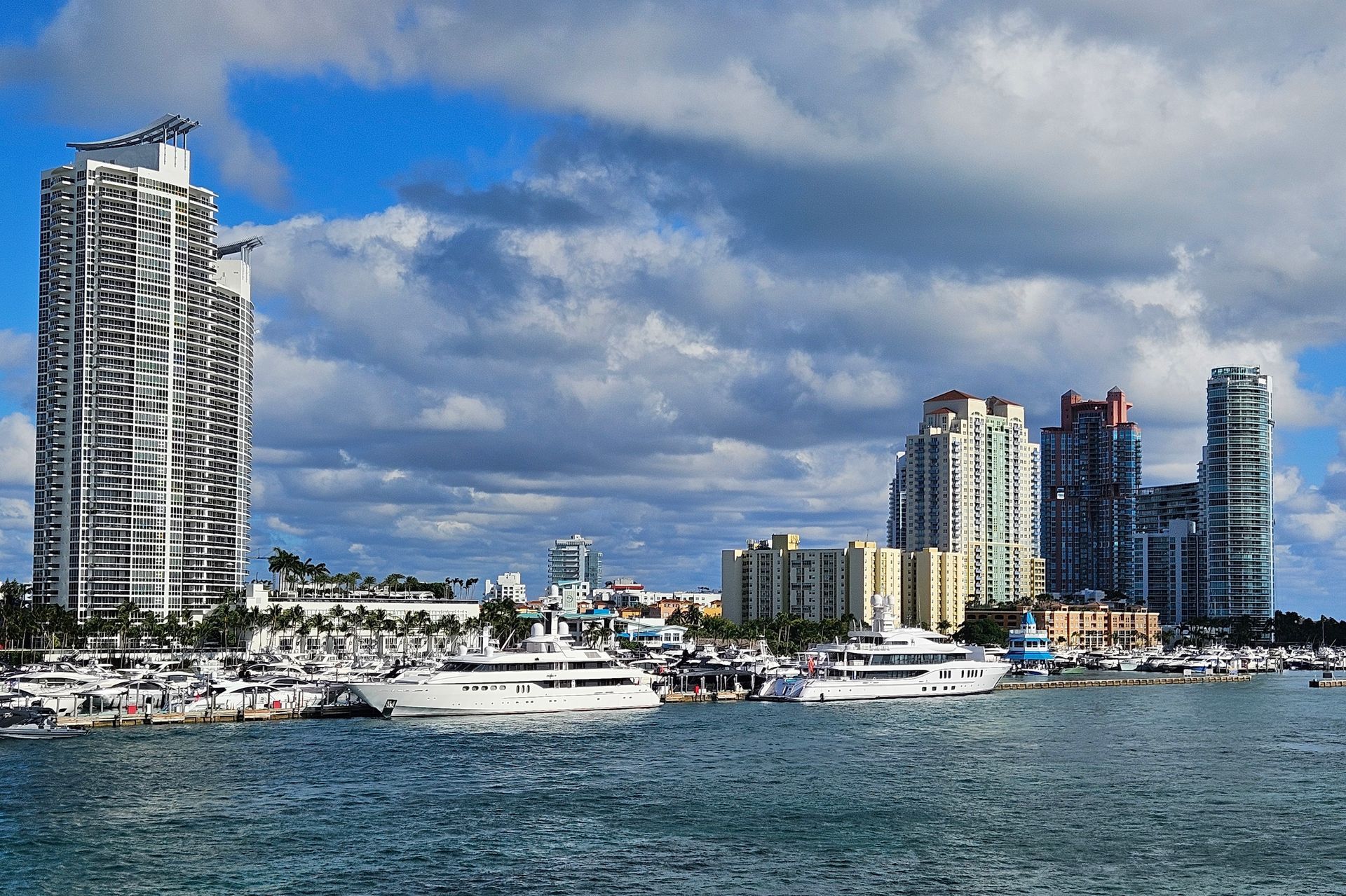 Skyline of Miami Beach with luxury yachts docked in front of tall buildings under a partly cloudy sky.