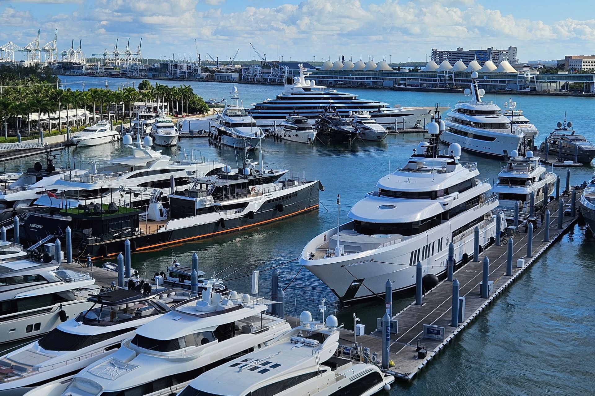 Luxurious yachts docked at a harbor, with a city skyline in the background. Blue water and sky, sunny day.