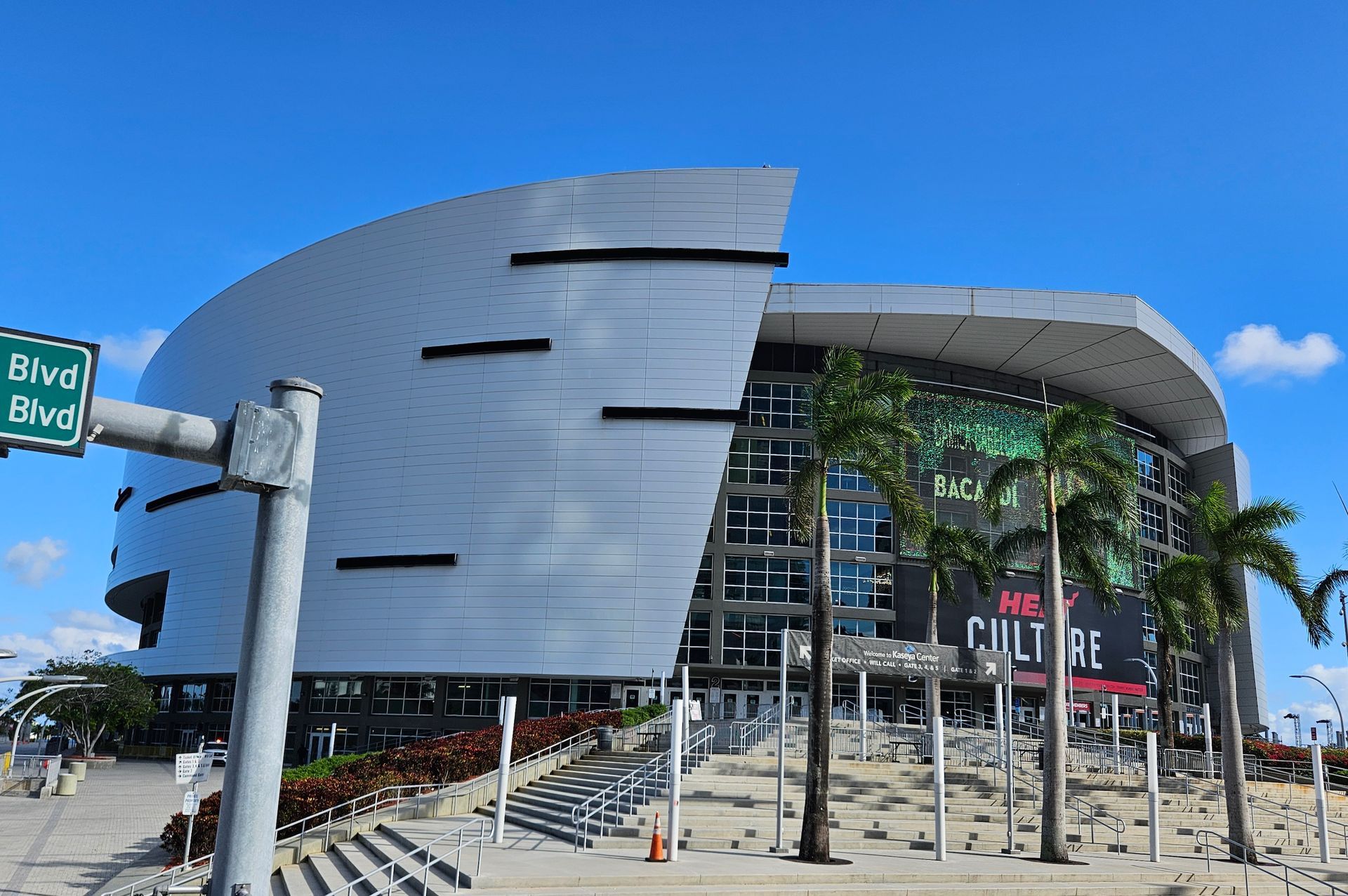 Miami's Kaseya Center, a modern arena with a curved metallic facade, palm trees, and a street sign.
