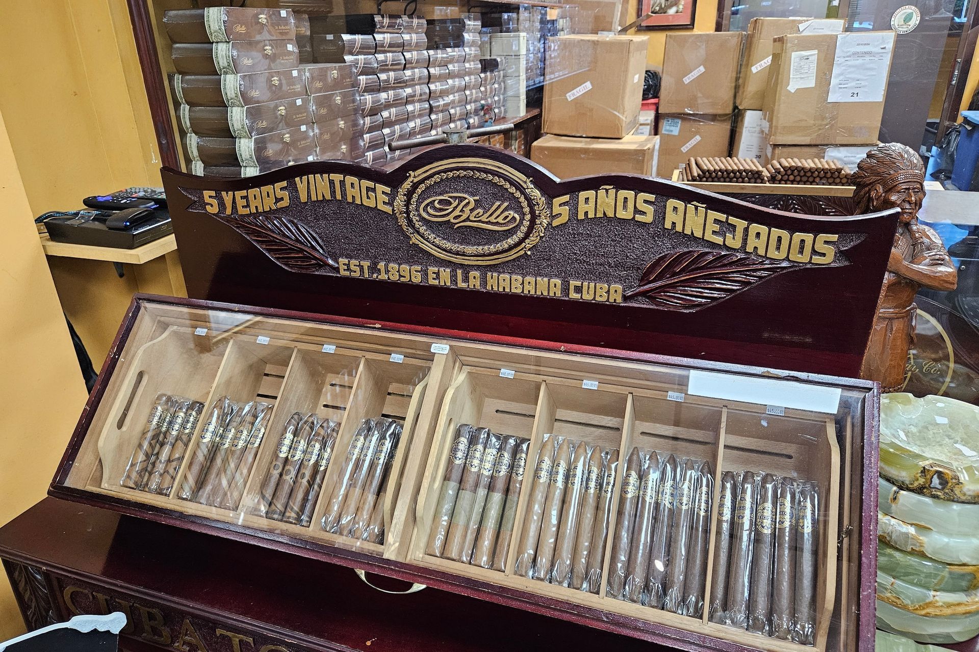 Cigar display case with rows of cigars, reddish-brown wood, with boxes in the background.