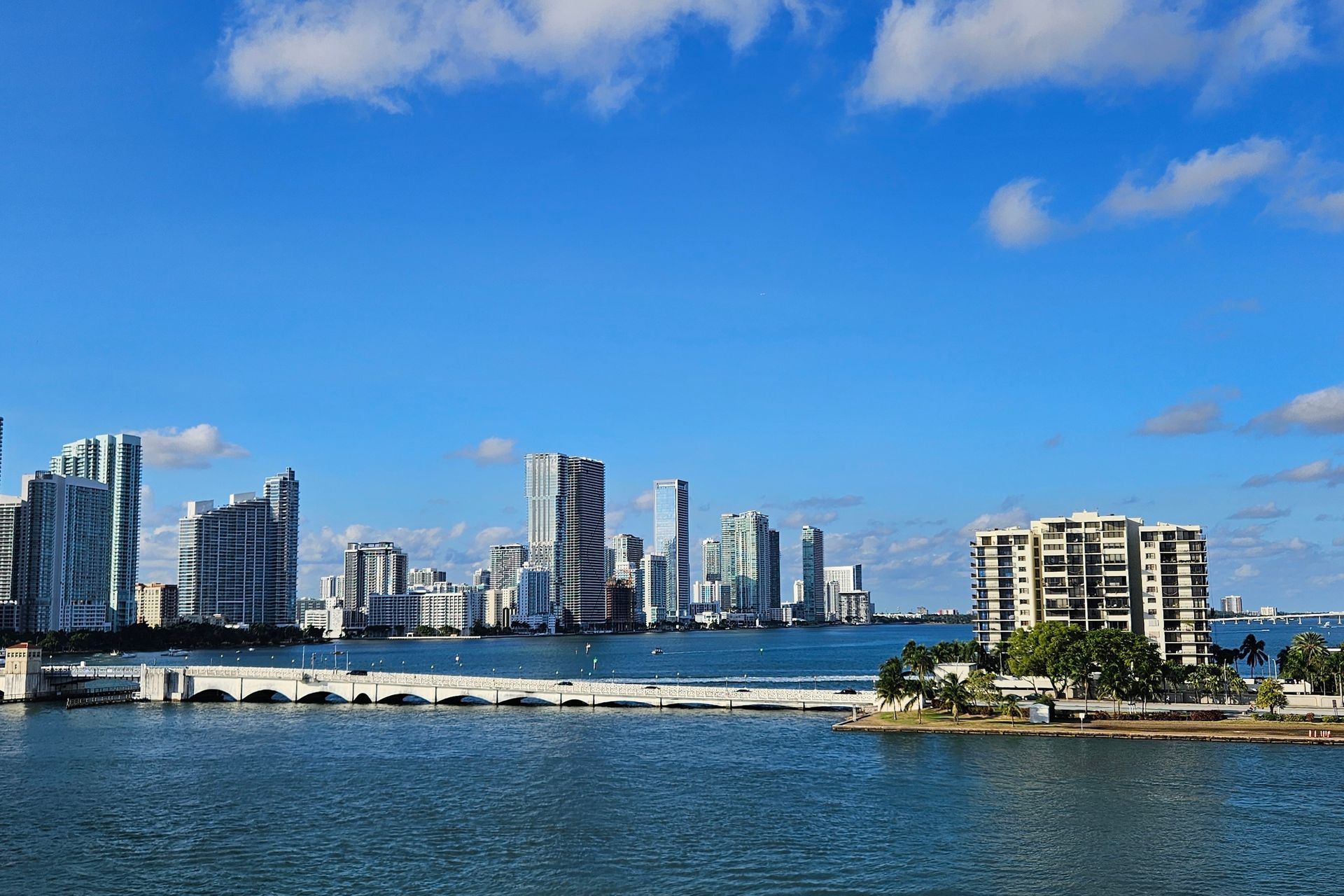 City skyline over water with a bridge, blue sky, and clouds.