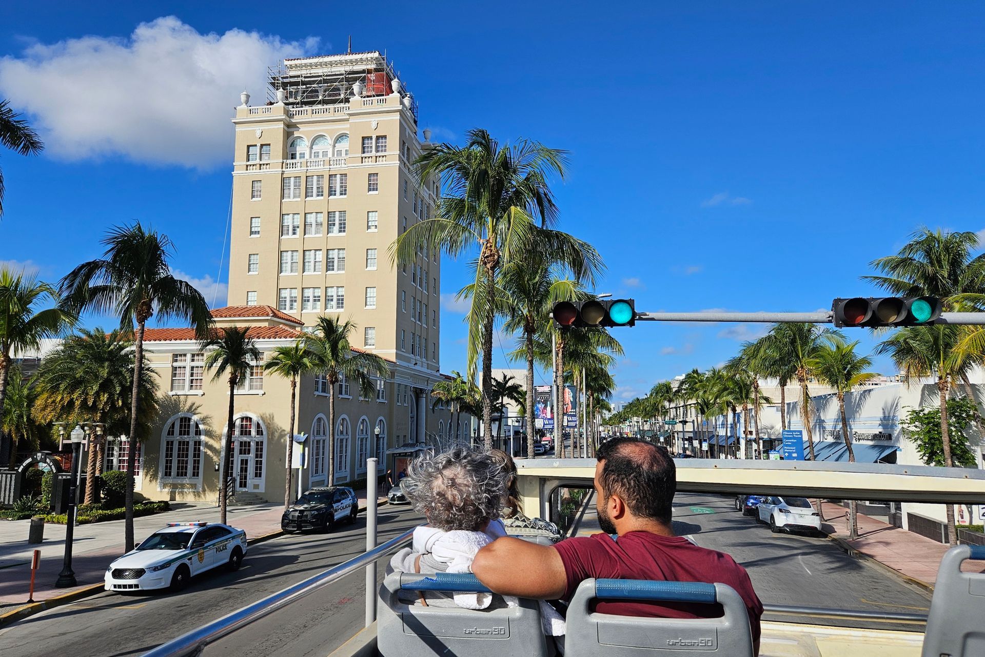 Tourists on a double-decker bus viewing a tall building with palm trees, under a blue sky in Miami.
