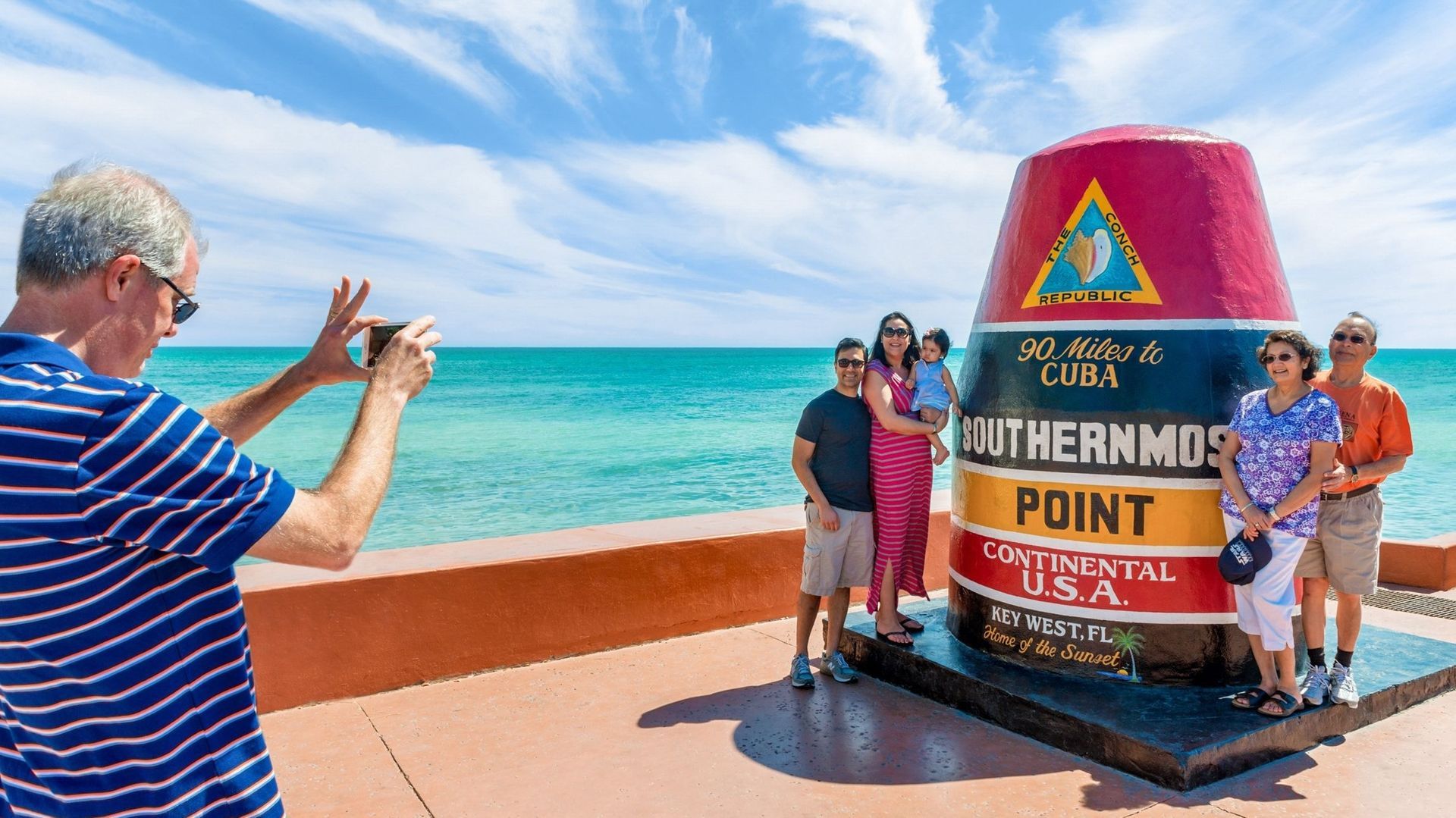 Man taking photo of a family posing by the Southernmost Point buoy in Key West, Florida; turquoise water and sky.