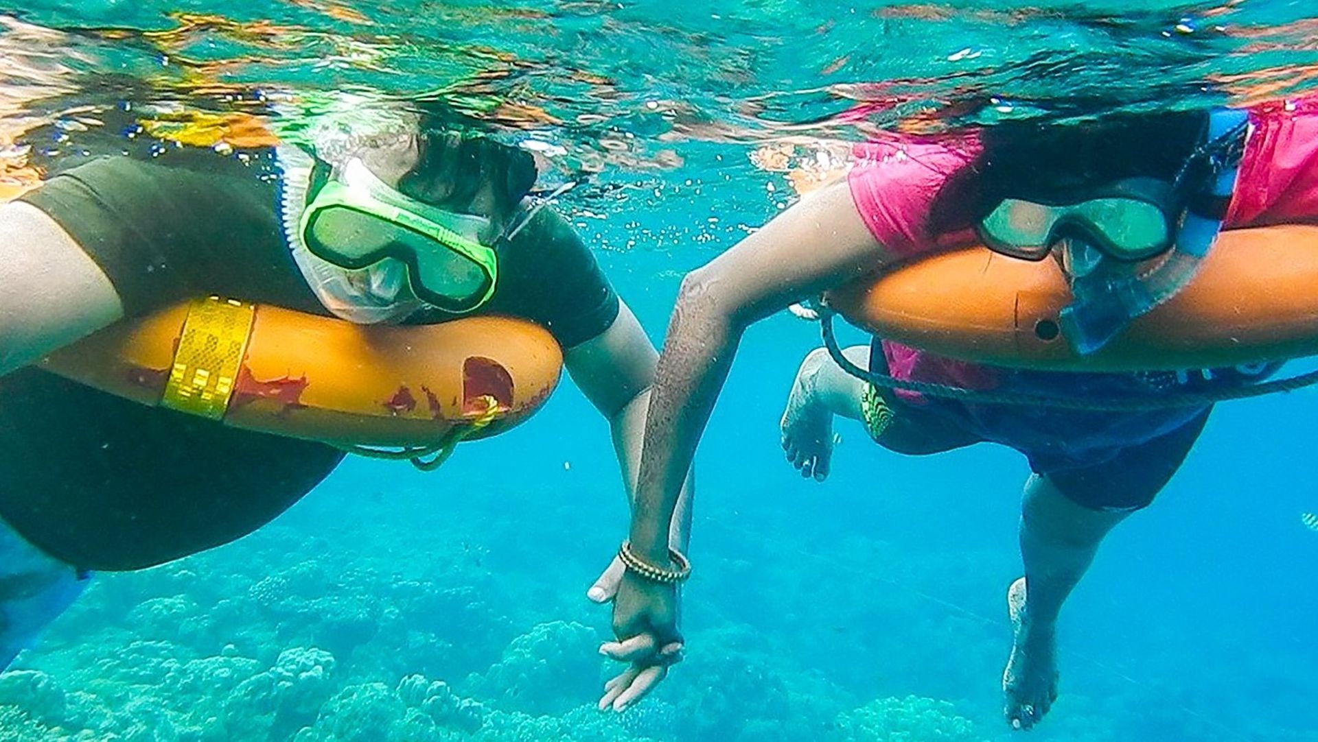 Two people snorkeling, holding hands underwater, using orange flotation devices.