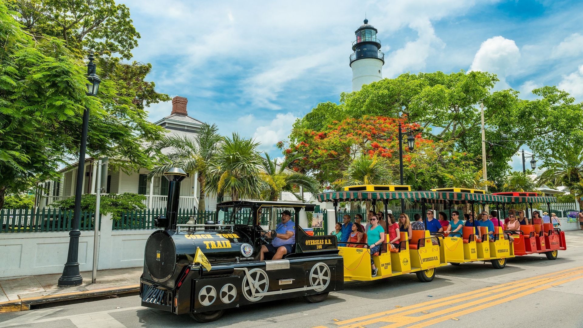 A black and yellow tour train drives along a street in Key West, Florida, past a lighthouse and historical buildings.