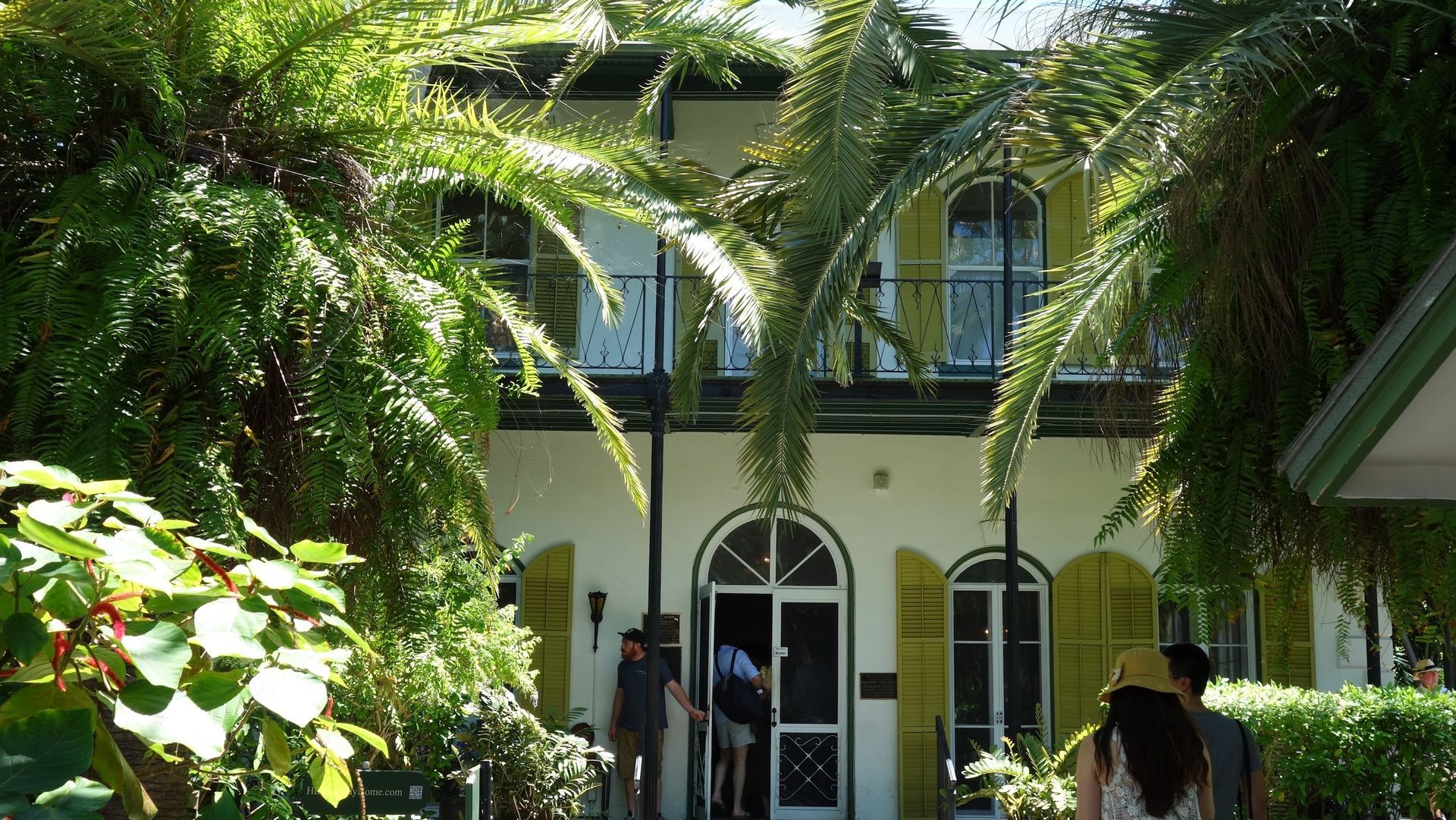 A white two-story house in Key West, Florida, with yellow shutters, palm trees, and visitors entering.