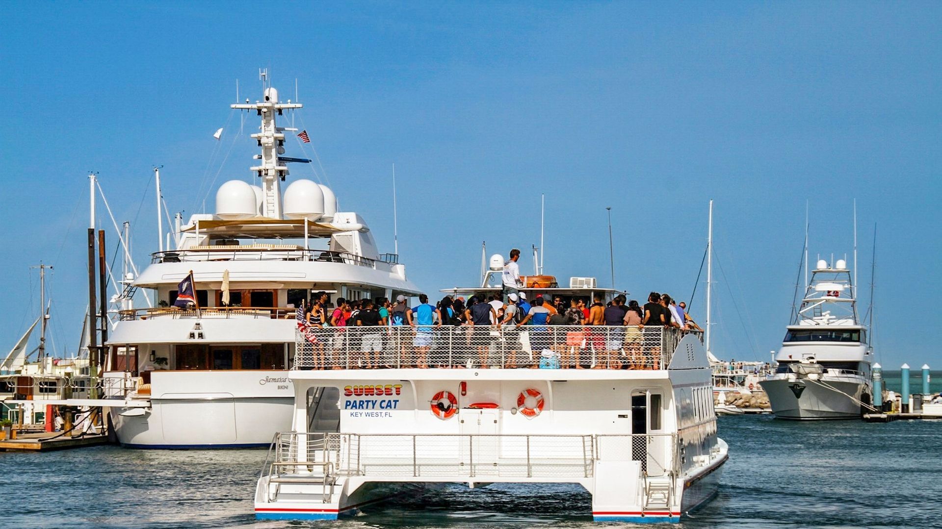 Boats docked in harbor: large white yacht, catamaran with people, fishing boat, blue sky.