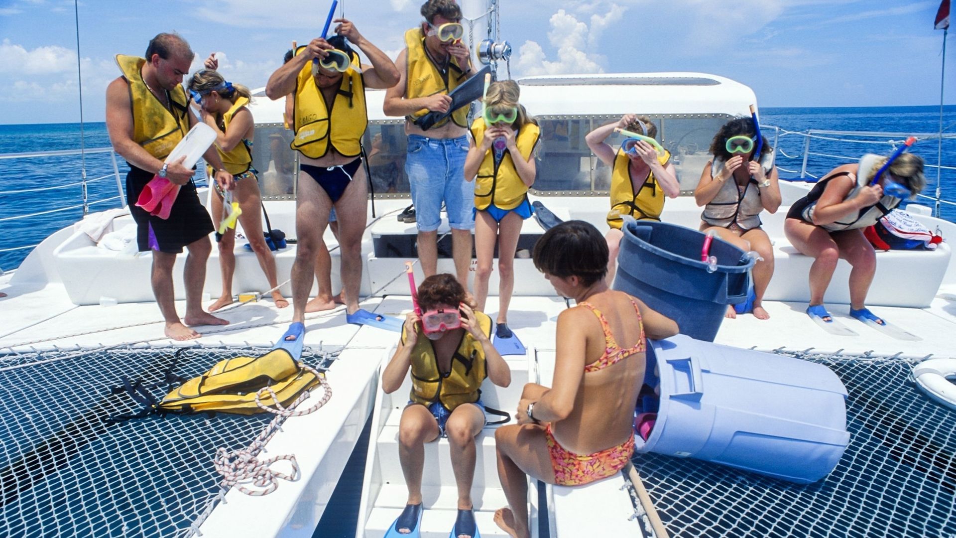 People on a catamaran preparing to snorkel: wearing life vests and goggles, by the ocean.
