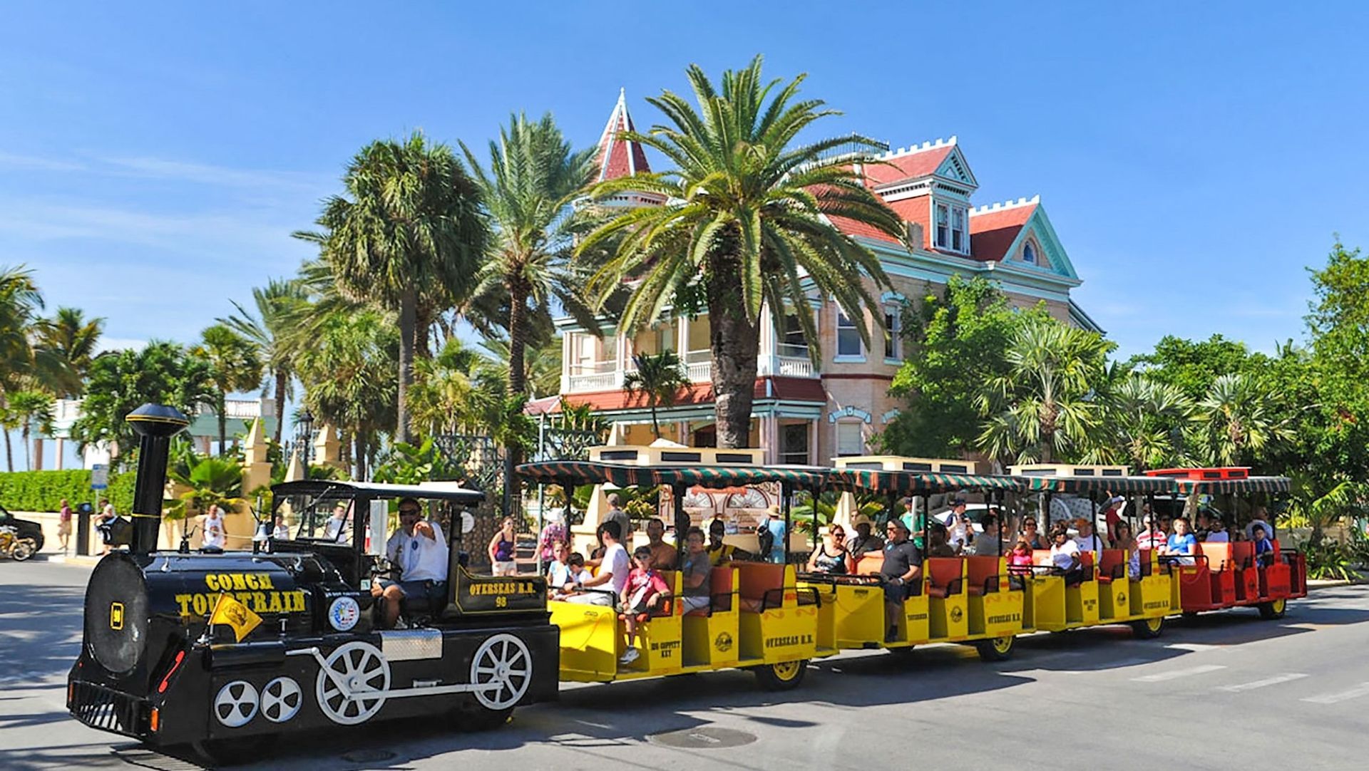 A tourist train in Key West, Florida, with passengers. The train is yellow and black, in front of a colorful Victorian house.