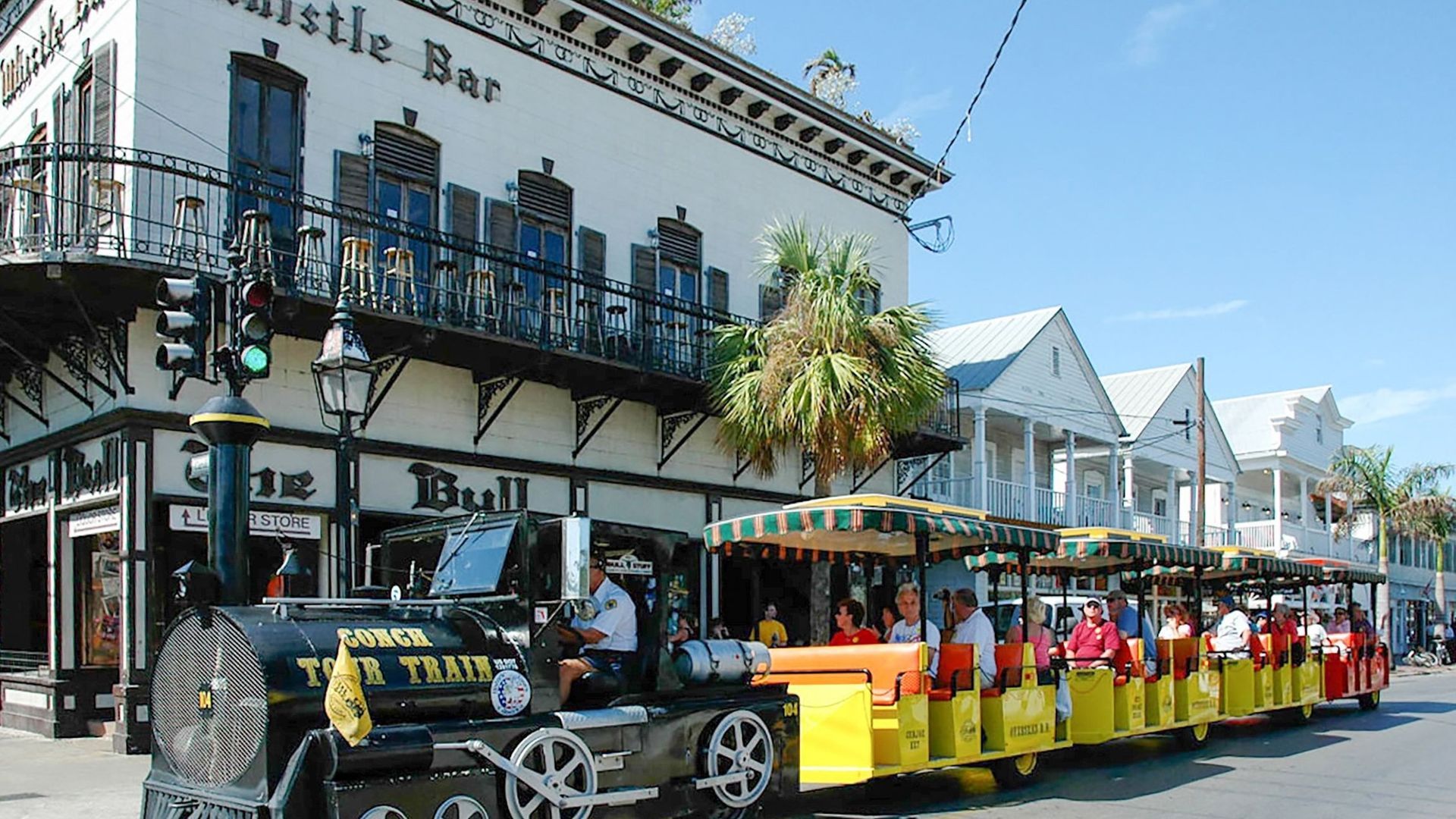 Key West Conch Train travels past a two-story building with a white exterior and a black and green bar.