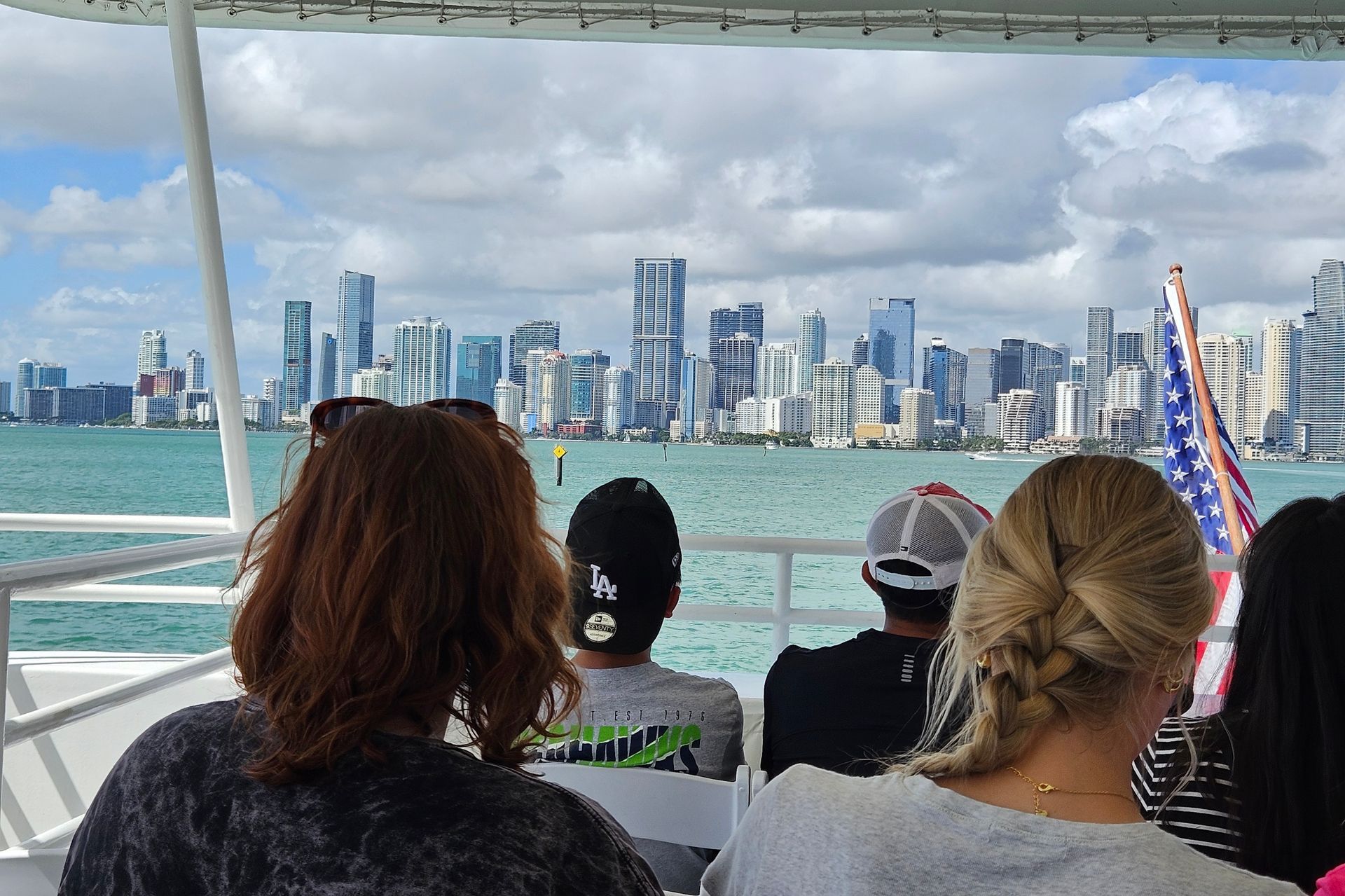 People on a boat looking at a city skyline with blue water and sky; American flag on the right.