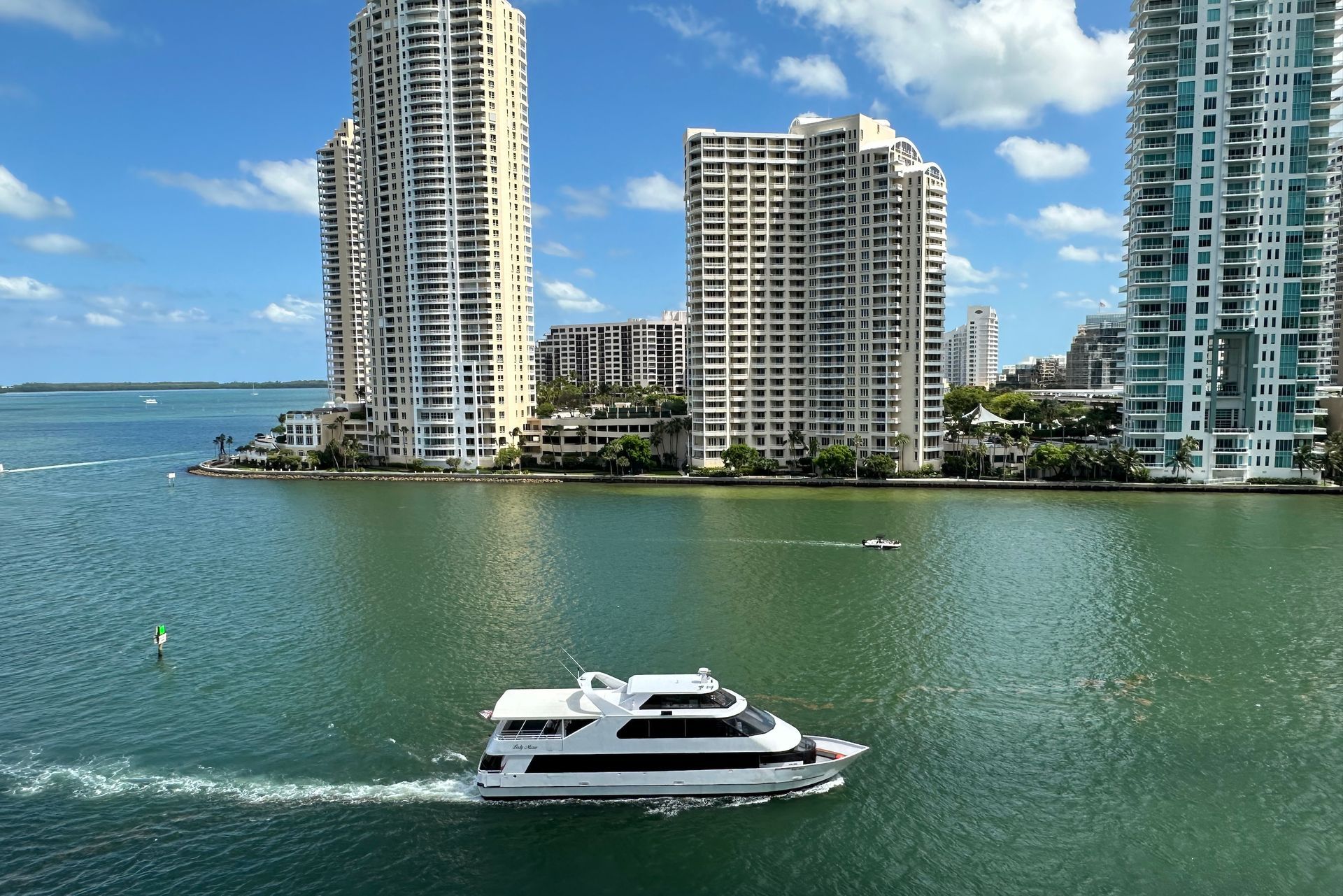 A white boat cruises on blue water, with tall buildings in the background under a blue sky.