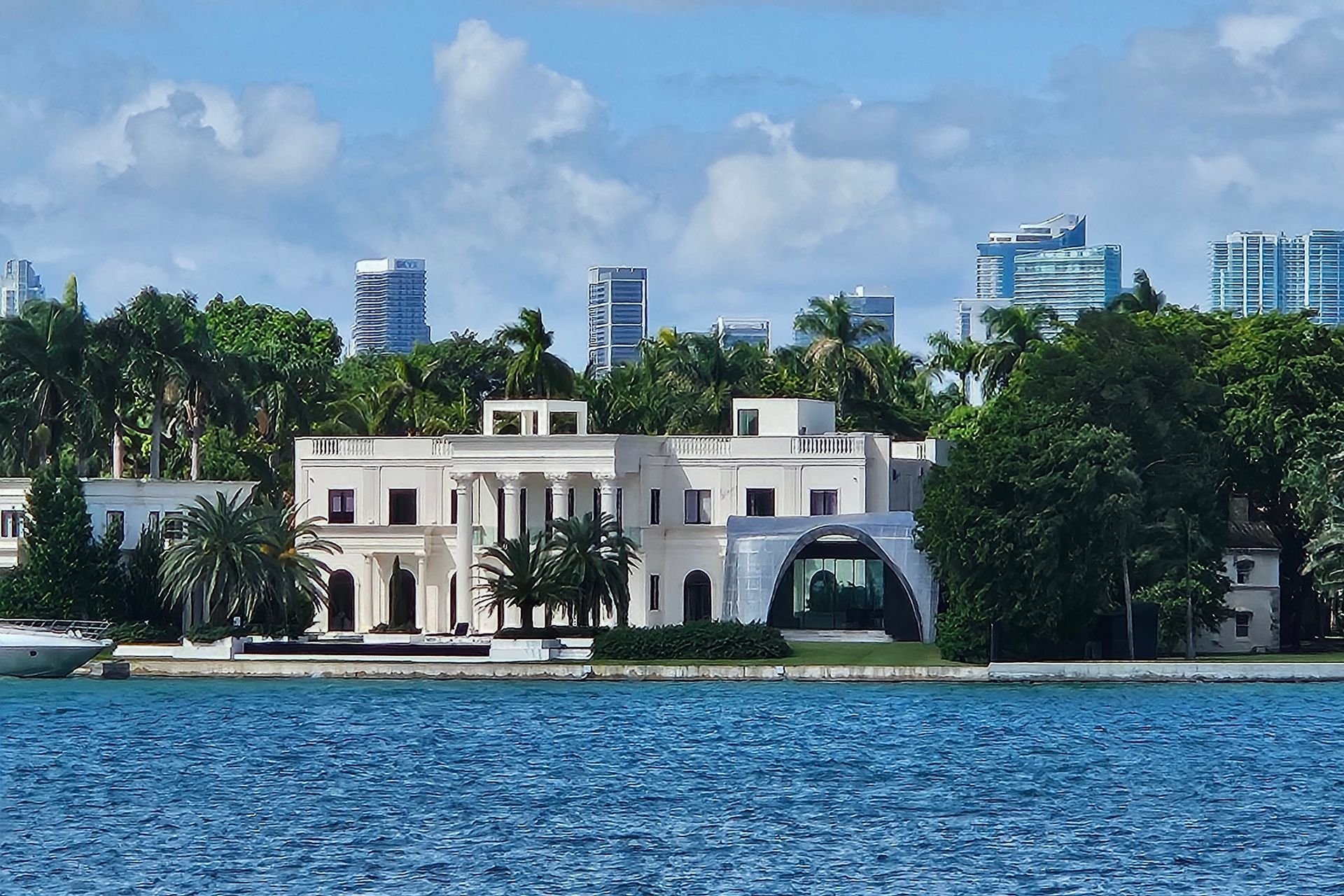 Mansion on water with city skyline in the background; blue water and sky, green trees.