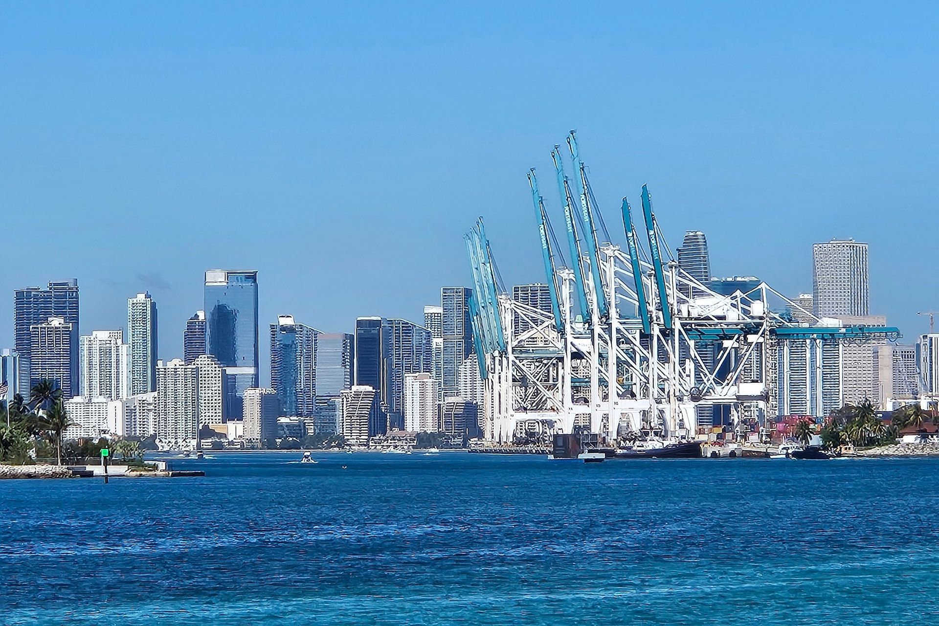 City skyline with tall buildings, blue water, and large cranes against a clear blue sky.