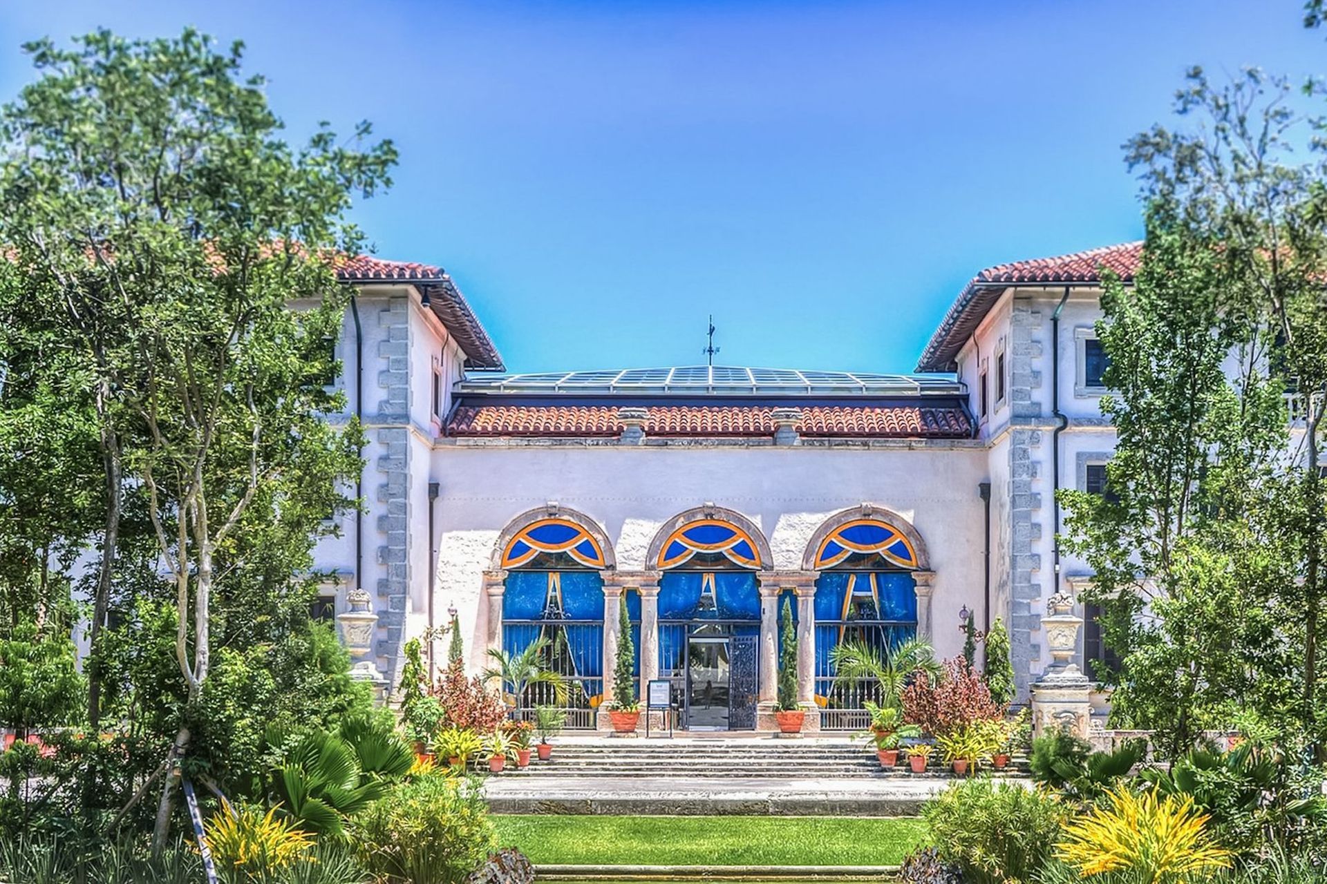 Mediterranean-style building with blue arched windows, red tile roof, and green landscaping under a bright blue sky.