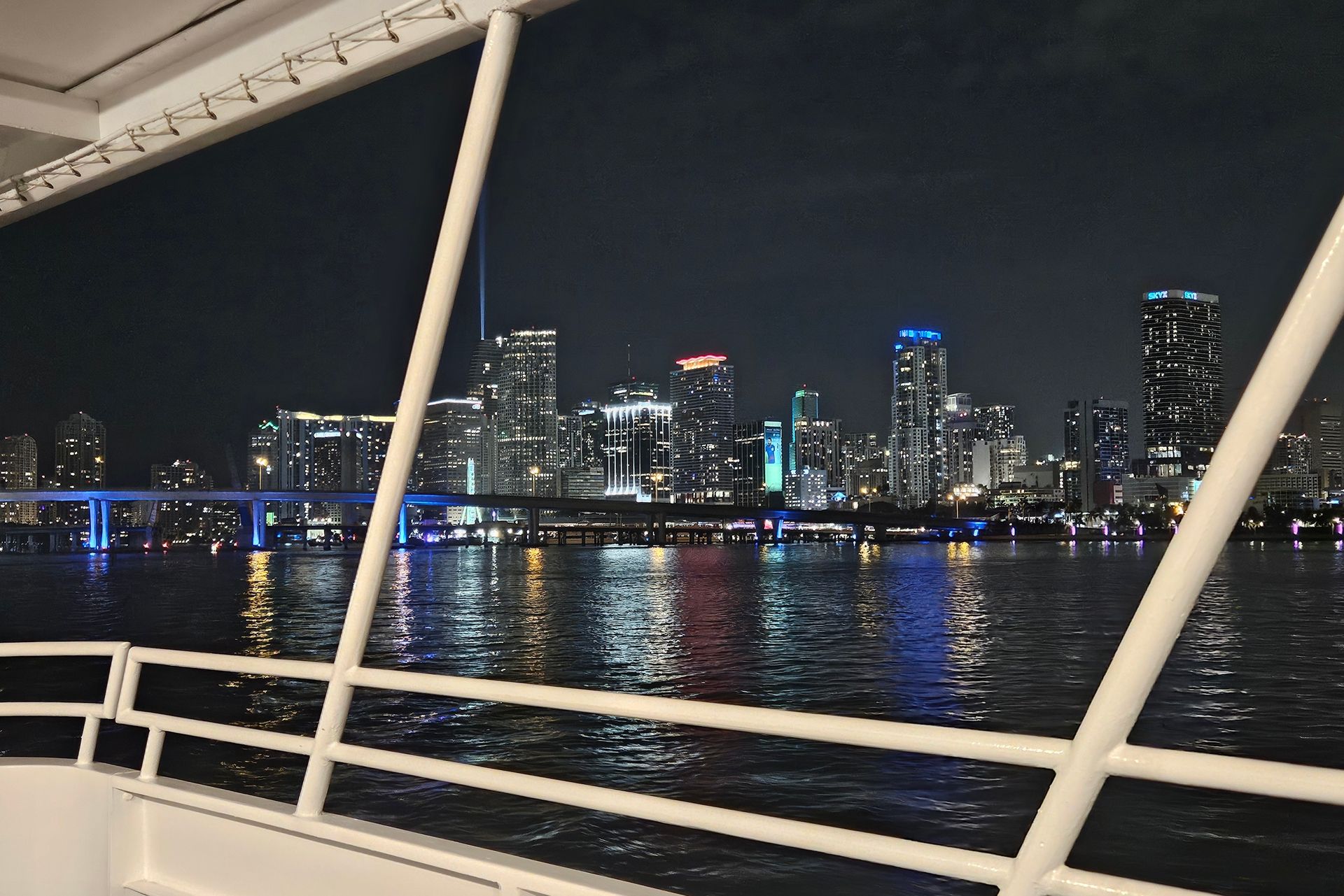 Night view of Miami skyline from a boat, lights reflected in water, white railings frame the view.