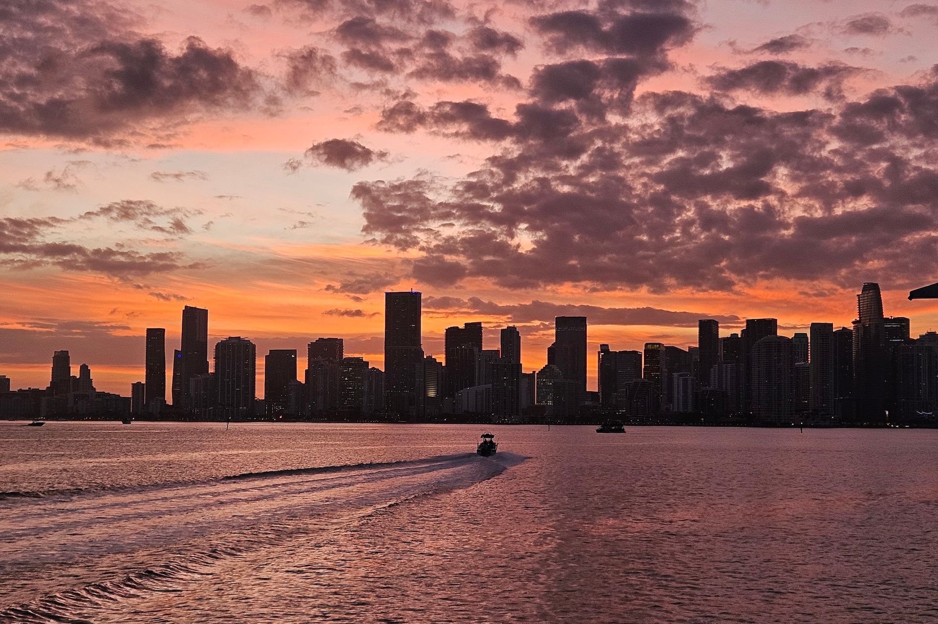 Sunset over city skyline, boat on water, orange and purple sky.