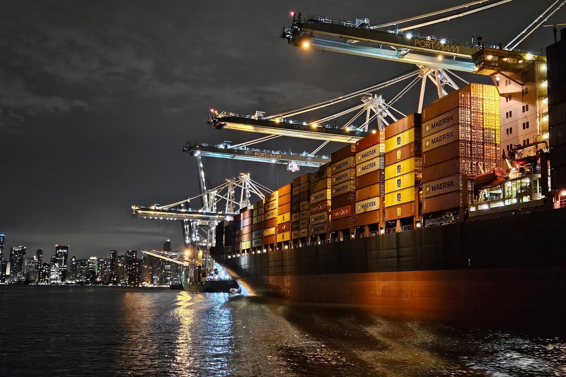 Cargo ship docked at a port, loading containers at night, illuminated by lights. City skyline in the background.