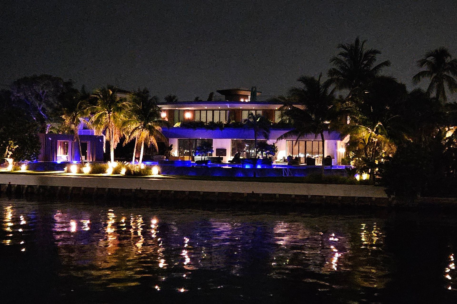 Luxury waterfront house at night, illuminated with blue and yellow lights reflecting in the water.