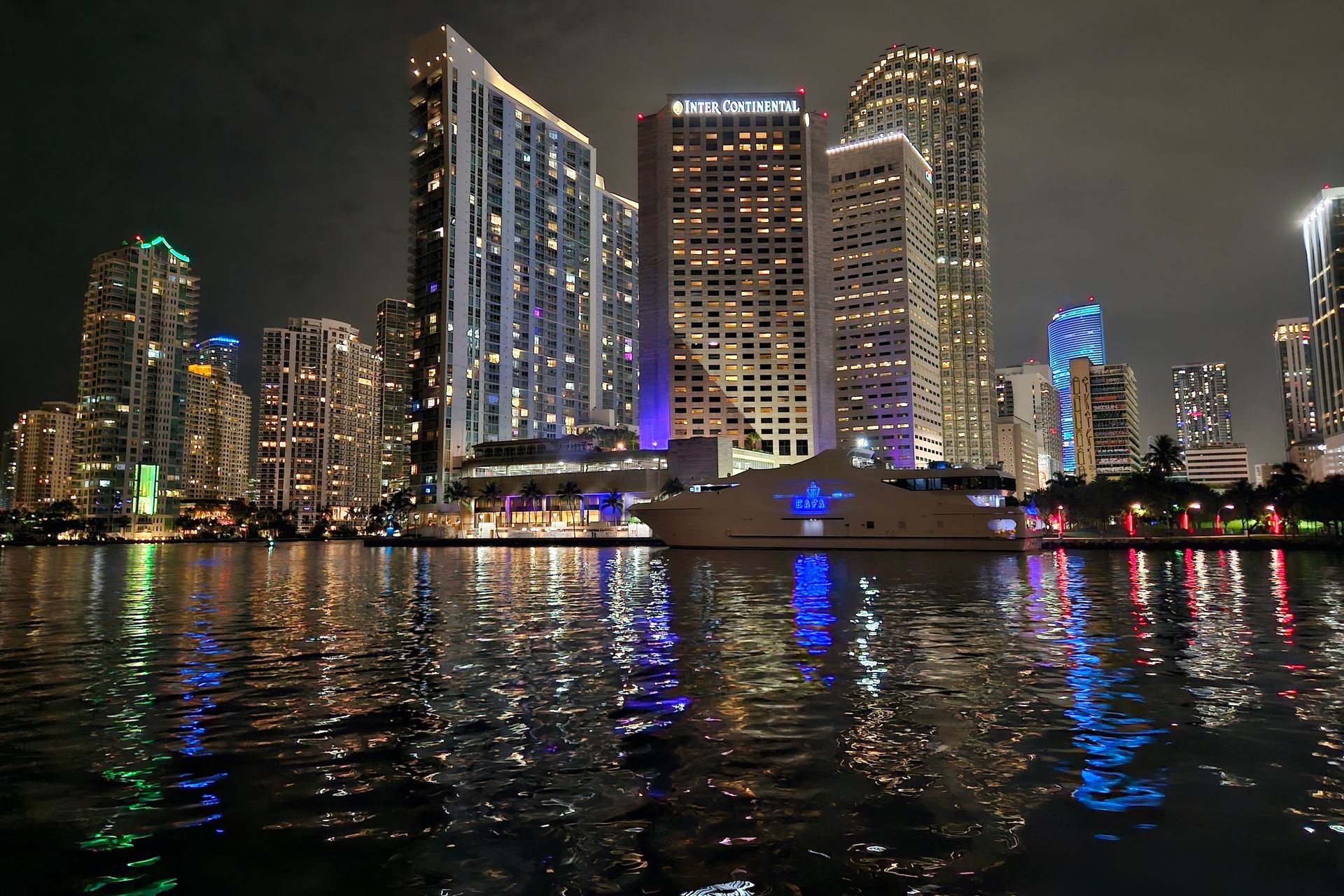 Nighttime cityscape with illuminated skyscrapers reflected in water; a yacht is in the foreground.