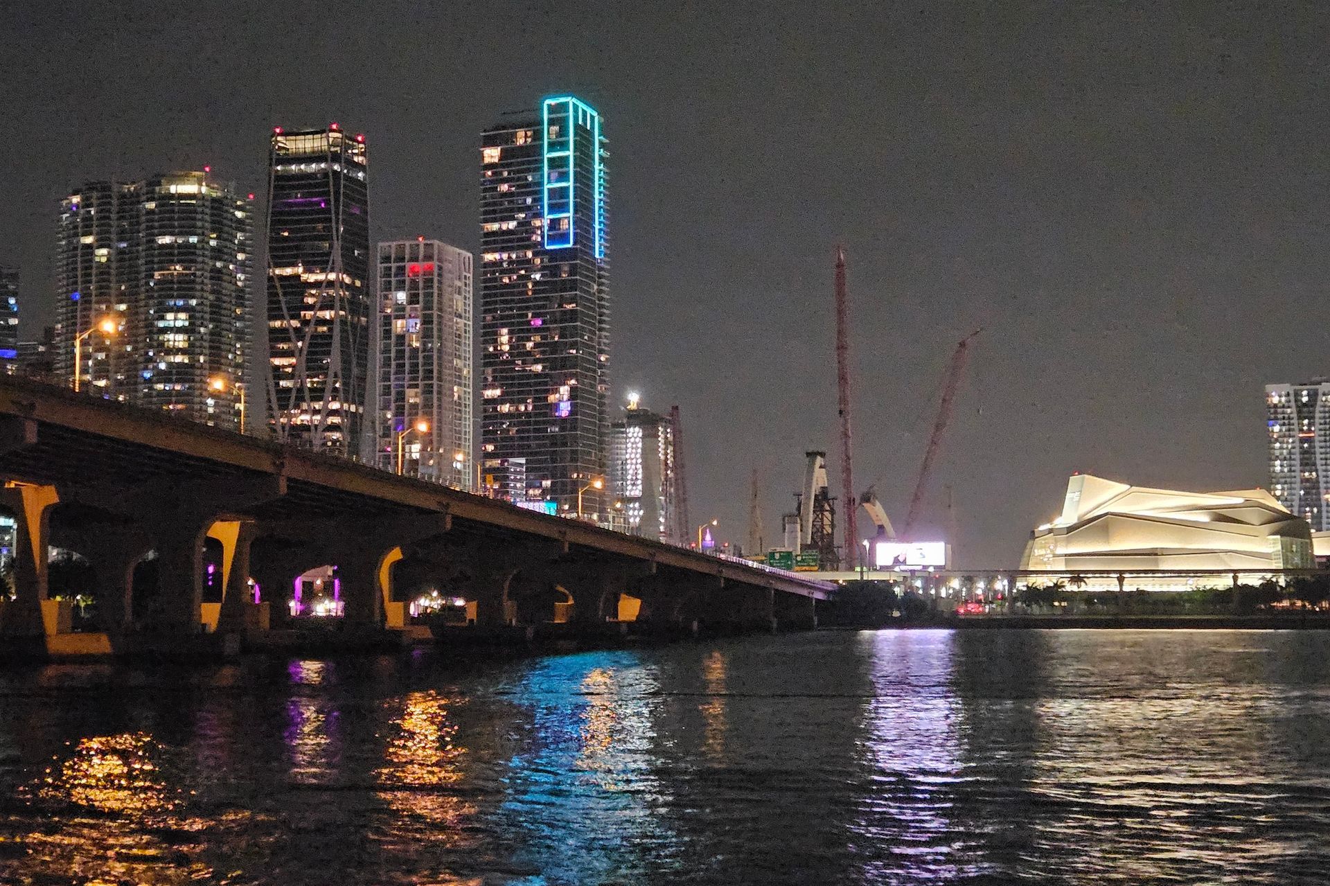 Nighttime cityscape with lit skyscrapers and a bridge over water.