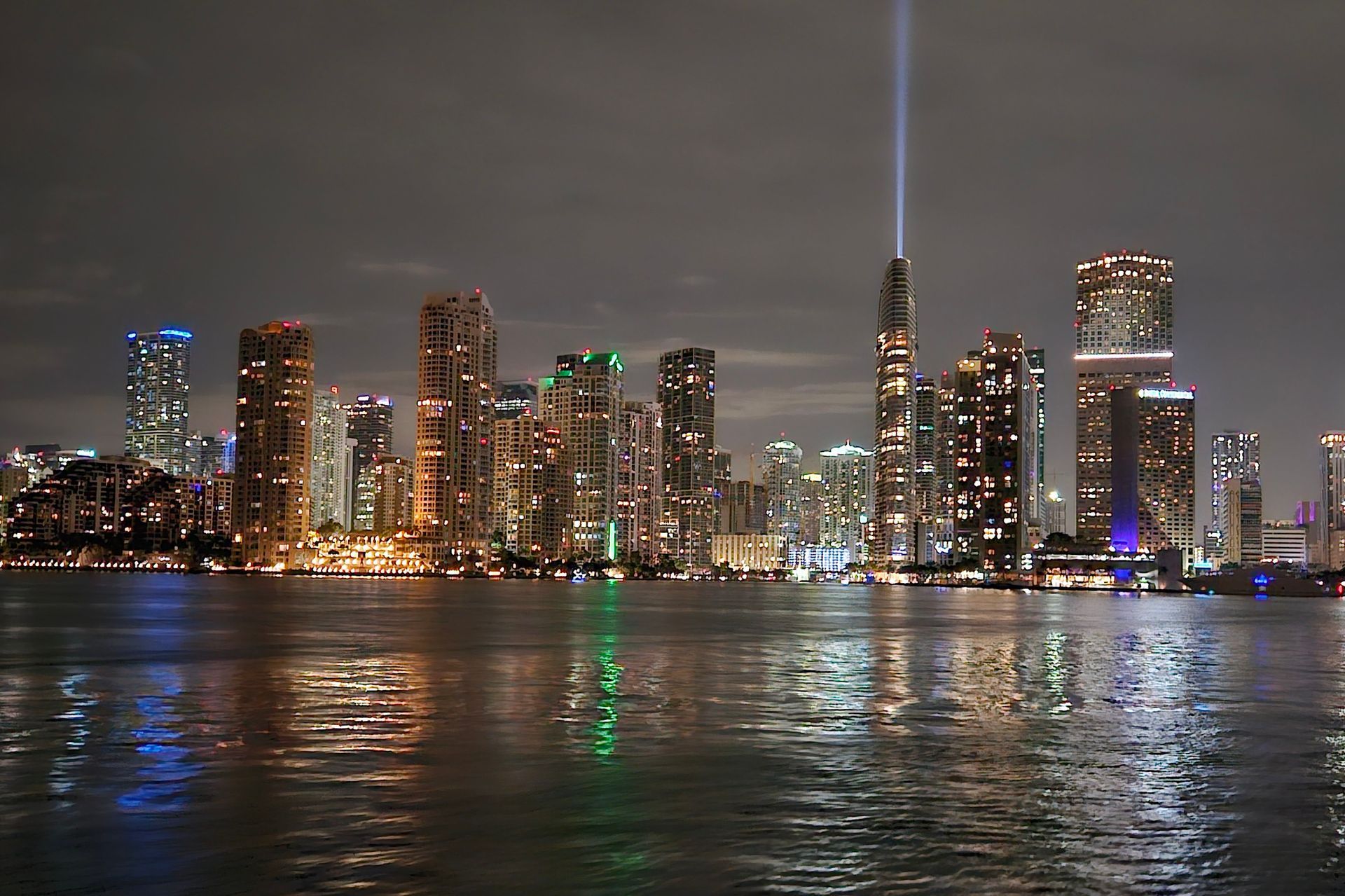 Nighttime skyline of a city with illuminated skyscrapers reflecting in the water. A bright light beam shines upwards.