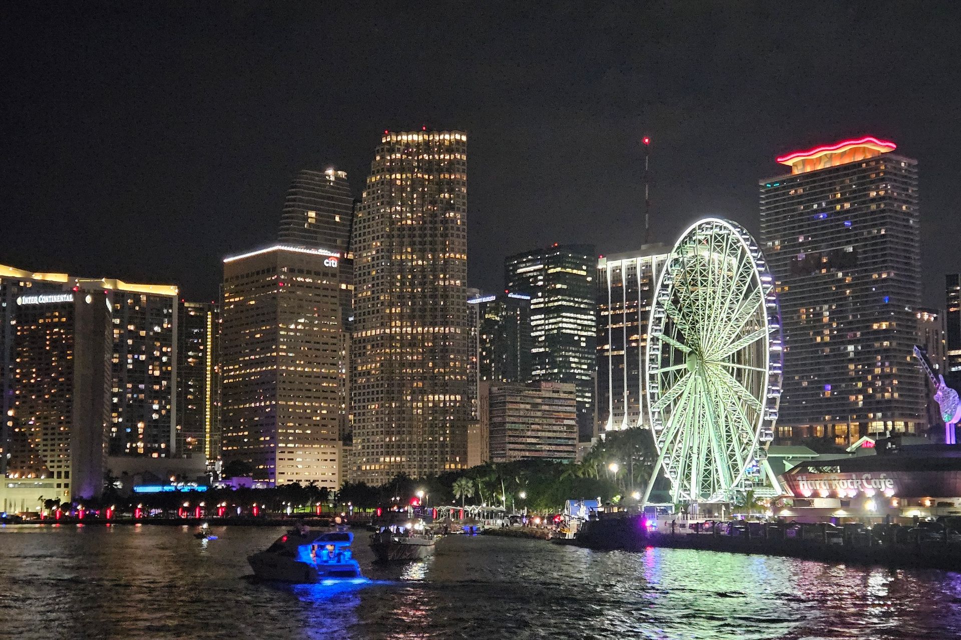 Night view of Miami skyline, illuminated skyscrapers, Ferris wheel, and boats on the water.