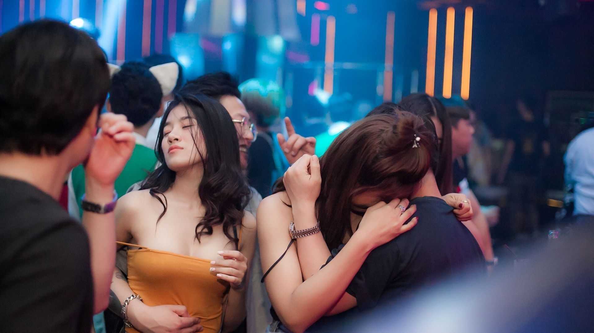 People dancing close together in a brightly lit club, with a woman in an orange dress.