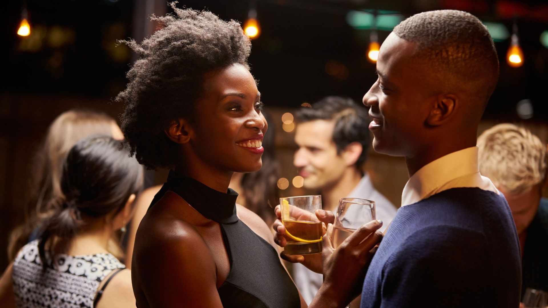 Smiling Black couple at a bar, holding drinks, engaged in conversation. Other people and dim lighting in background.
