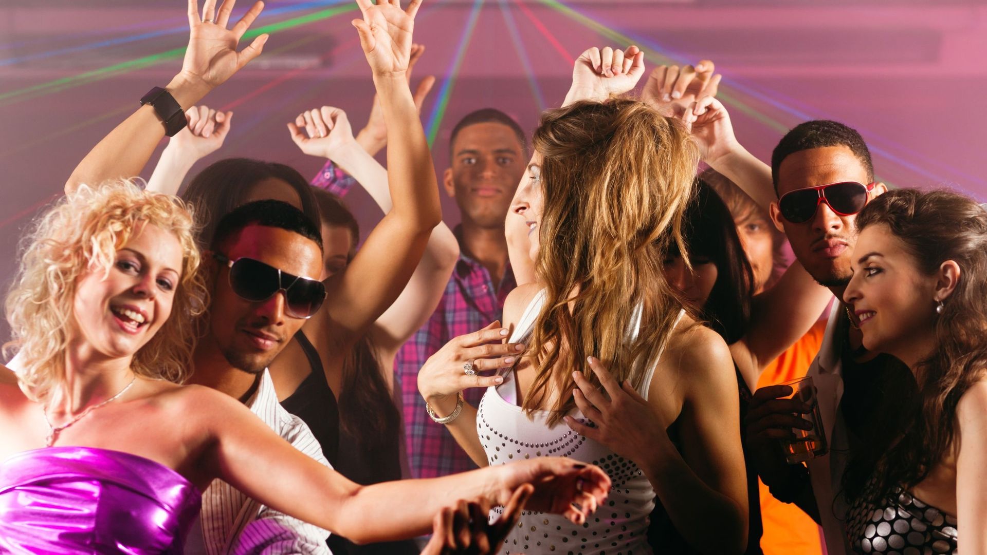 People dancing in a nightclub, arms raised, wearing casual and party attire. Lights and smiles.