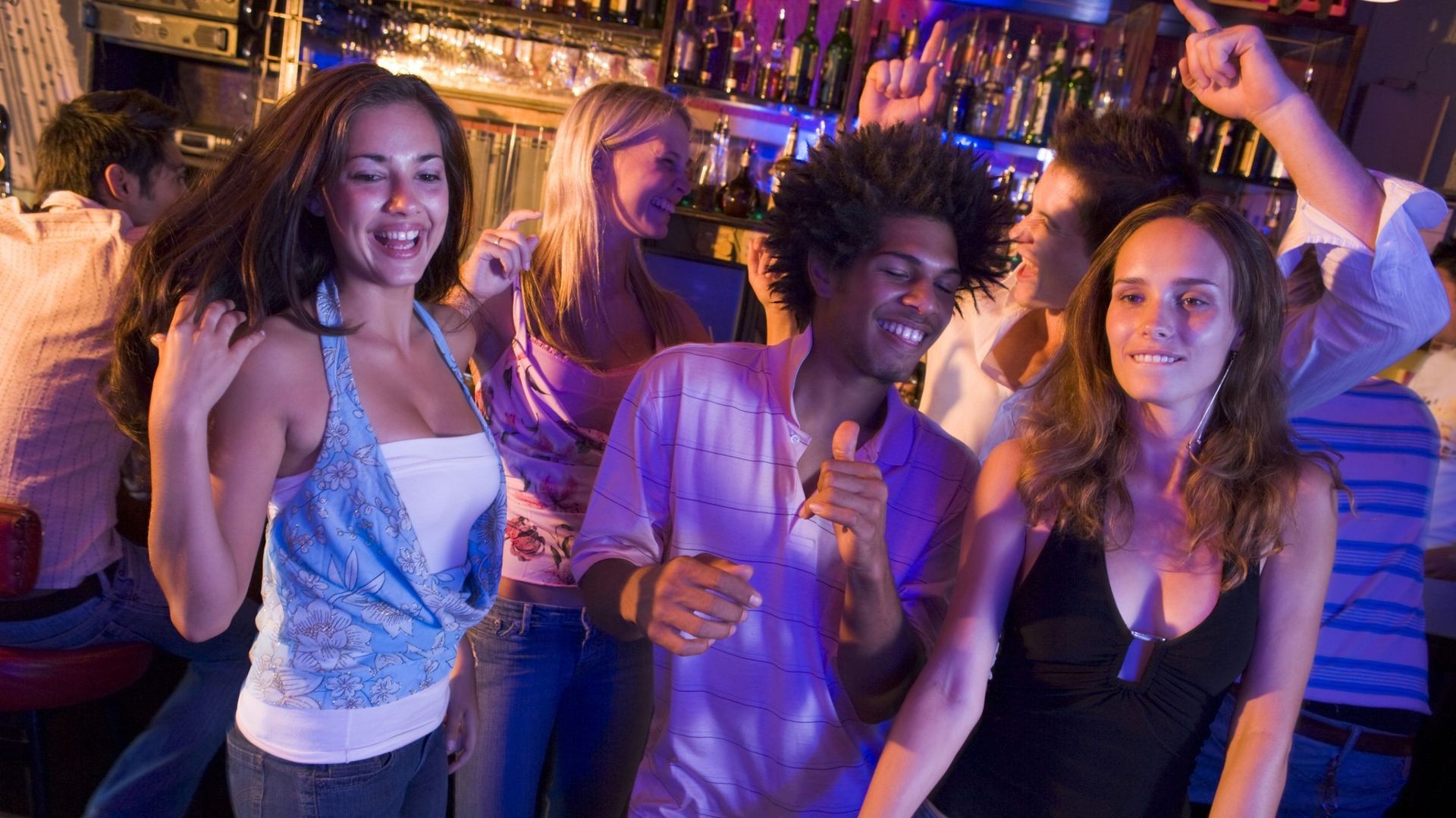 People dancing and smiling in a brightly lit bar with bottles visible behind the bar.