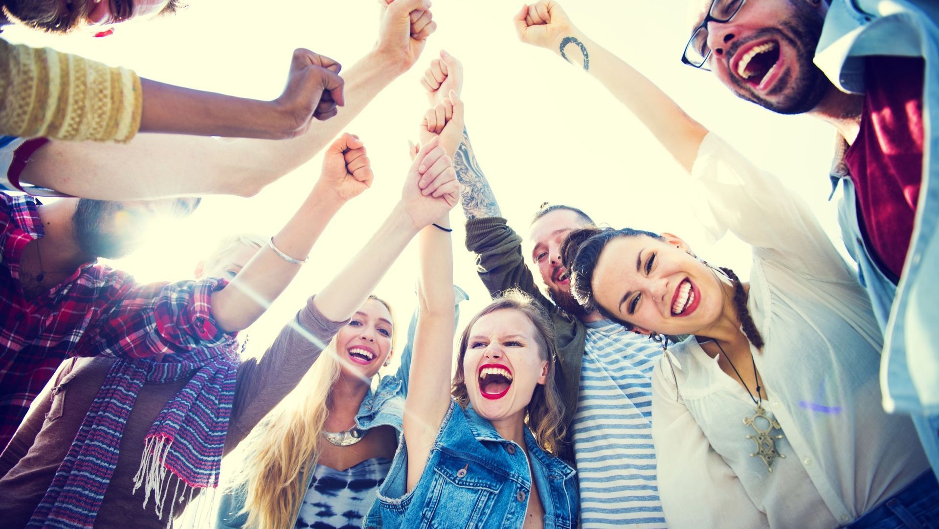 Group of diverse young adults celebrating together, arms raised in a circle, laughing and cheering.