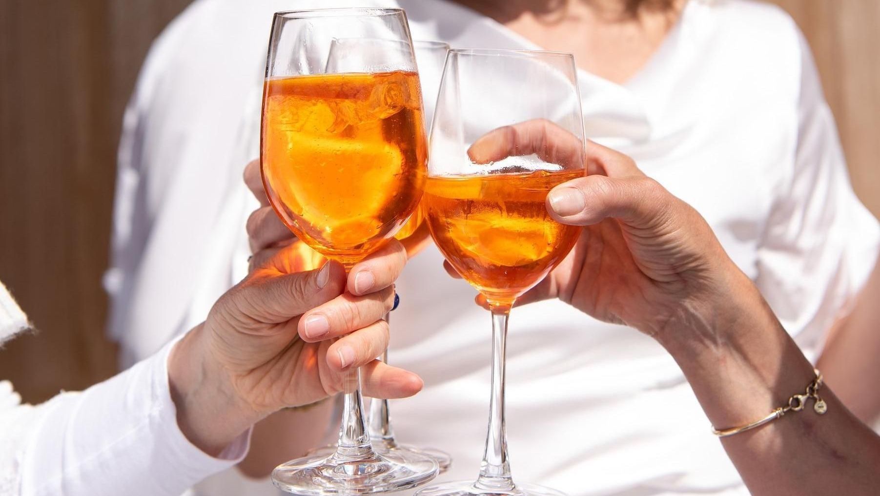 Two people in white clothing clink glasses of orange Aperol Spritz outdoors.