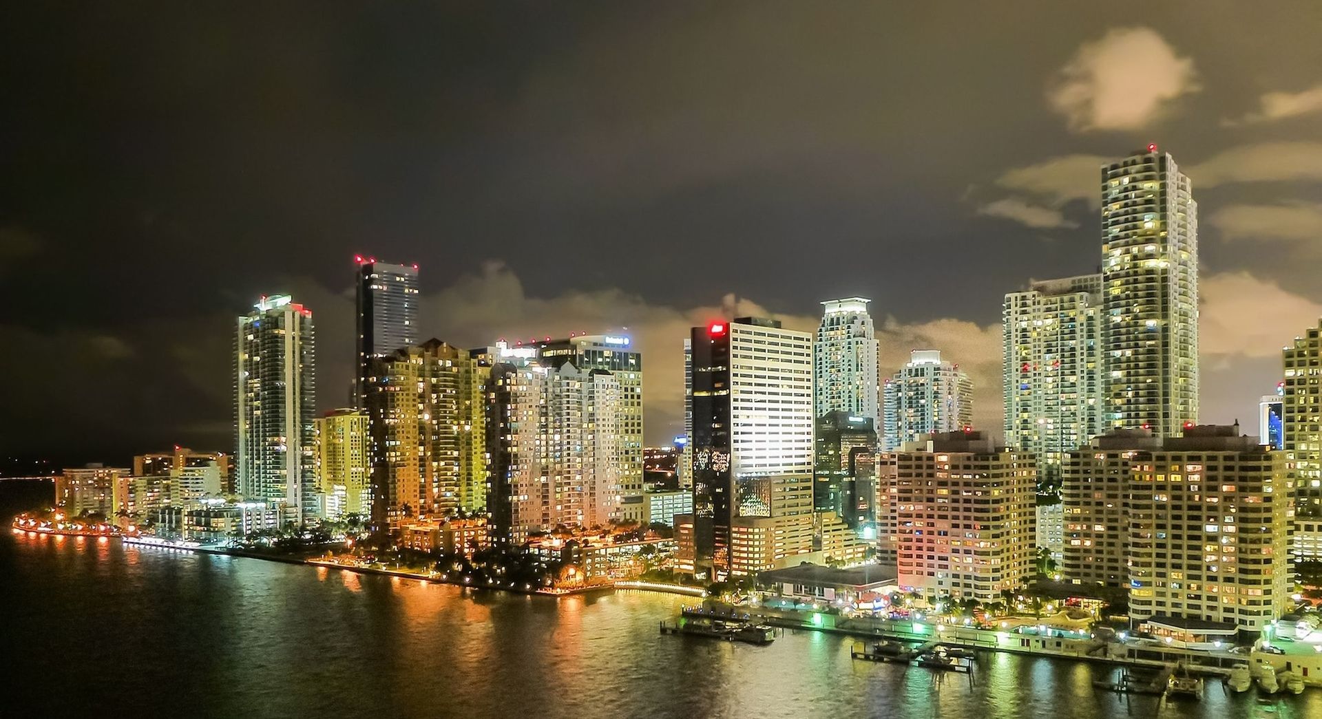 Nighttime skyline of Miami, Florida, with brightly lit skyscrapers reflecting in the water.