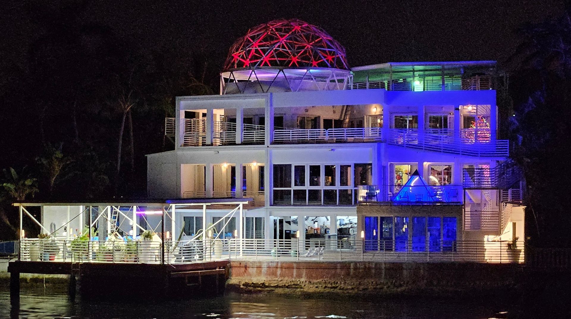Multi-story, white building with red-lit dome, illuminated blue and white, at night on the water.