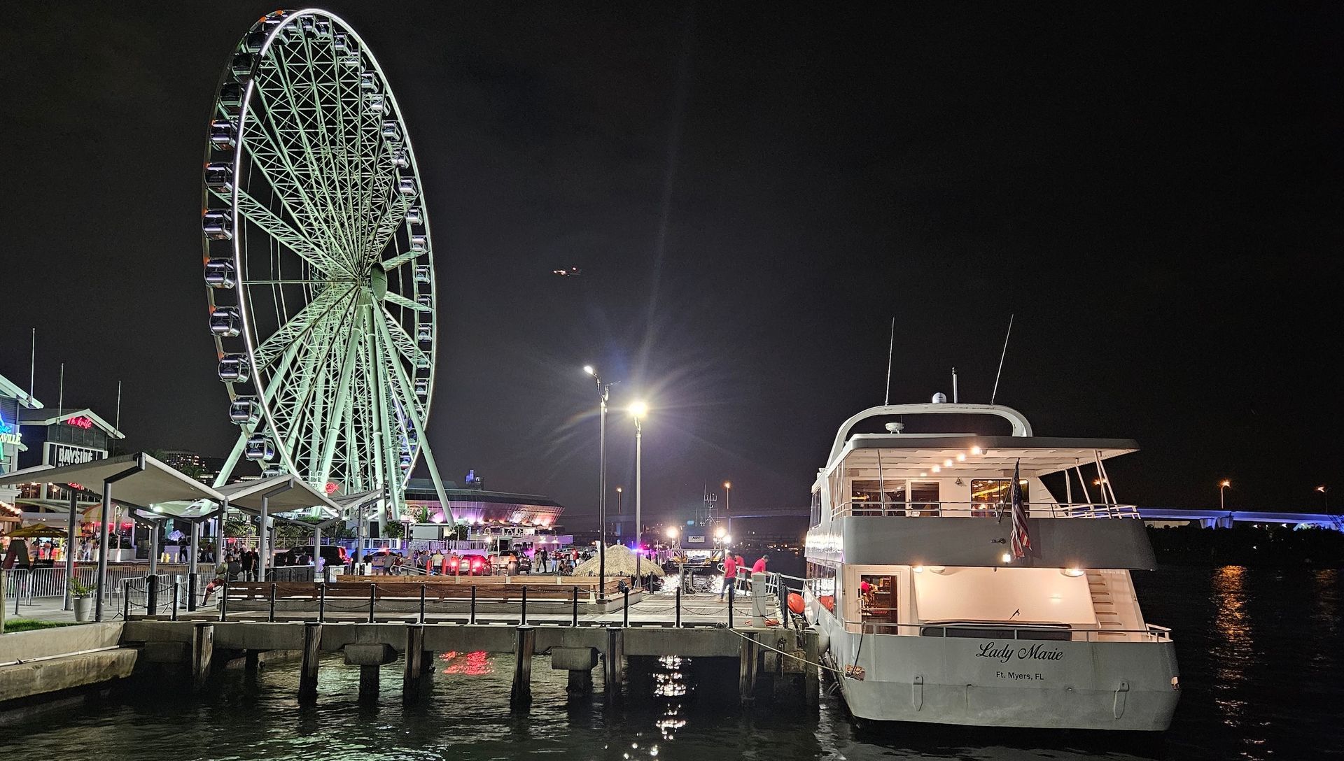 Night scene with lit Ferris wheel, pier, and docked yacht.