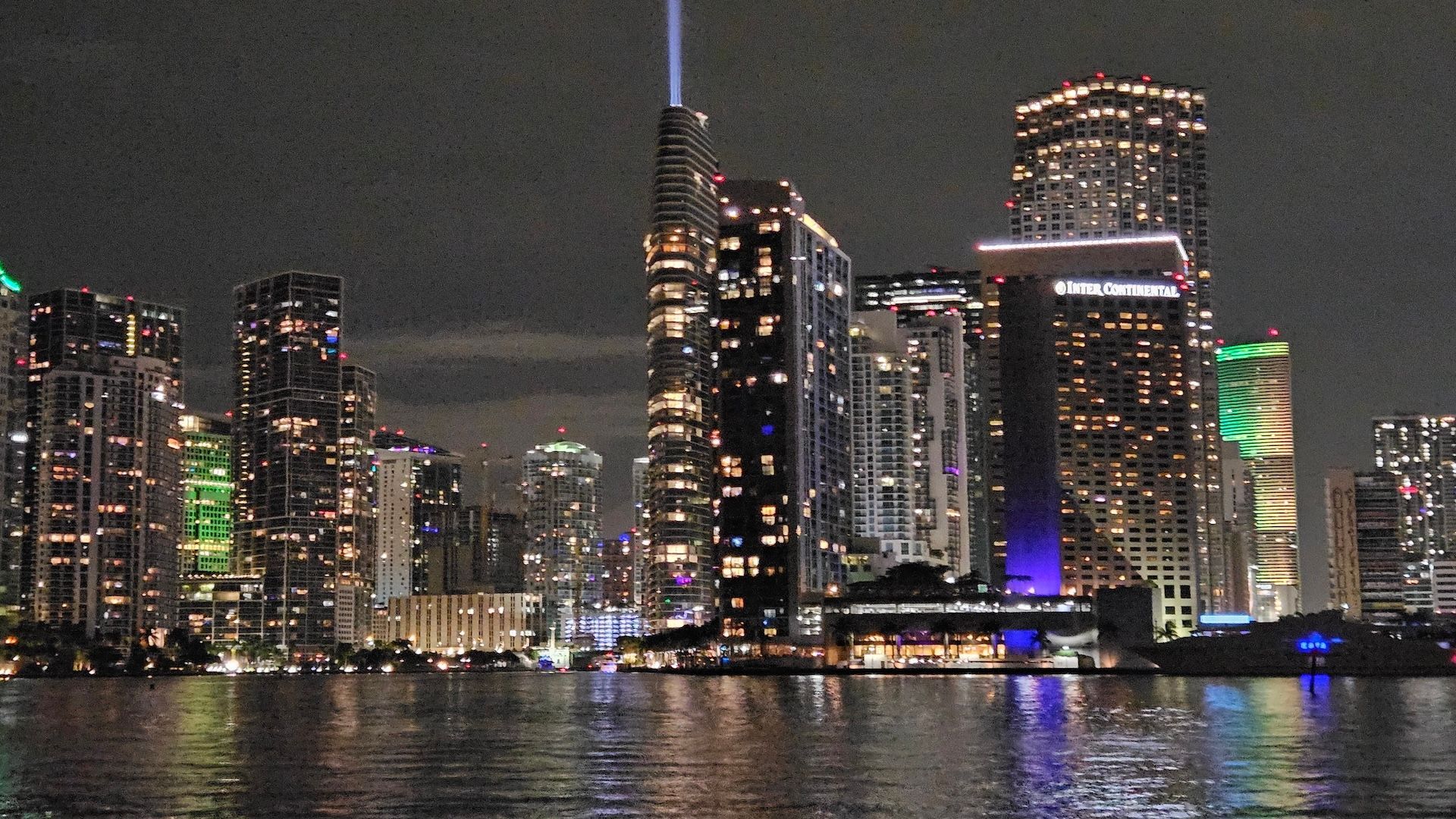 Night cityscape of Miami, lit buildings reflecting on dark water.