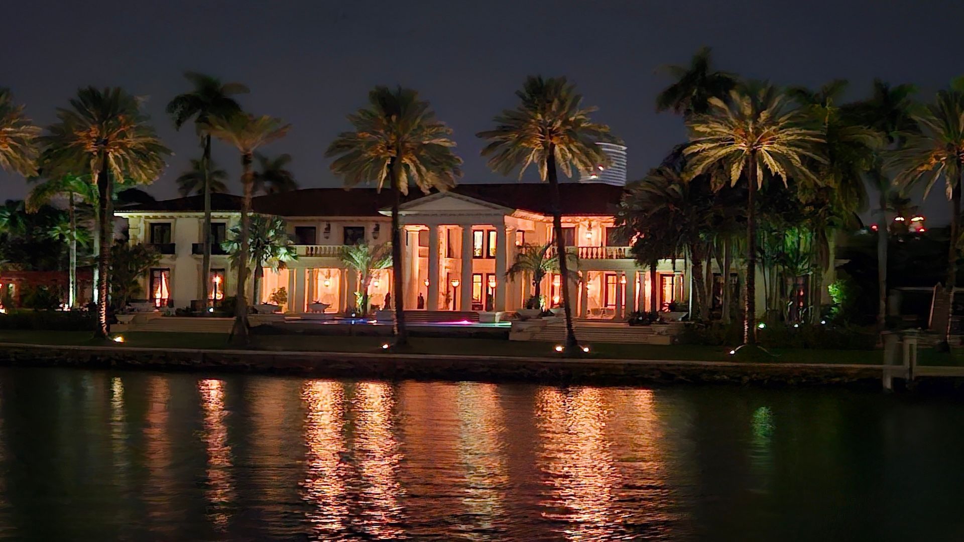 A lavish, illuminated mansion with palm trees, reflected in dark water at night.