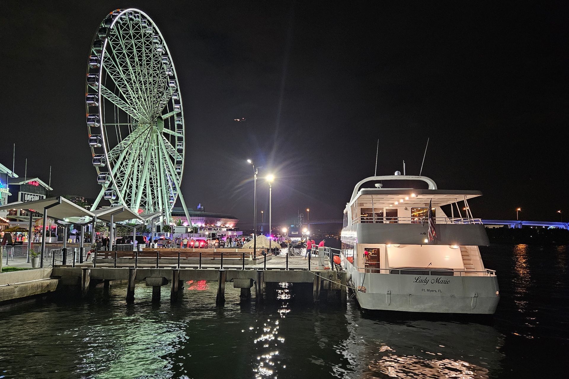 A nighttime view of a Ferris wheel and a docked yacht near a pier. The scene is illuminated.