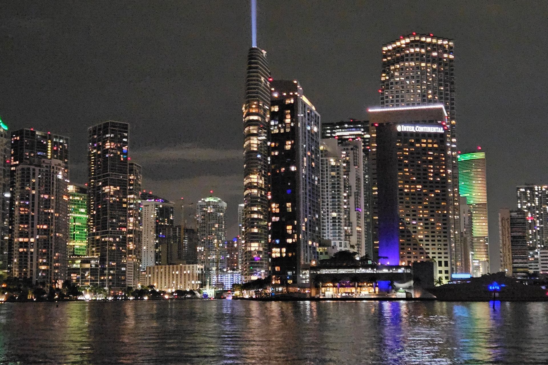 Miami skyline at night, illuminated skyscrapers reflect in the water, a vertical light beam streaks into the sky.